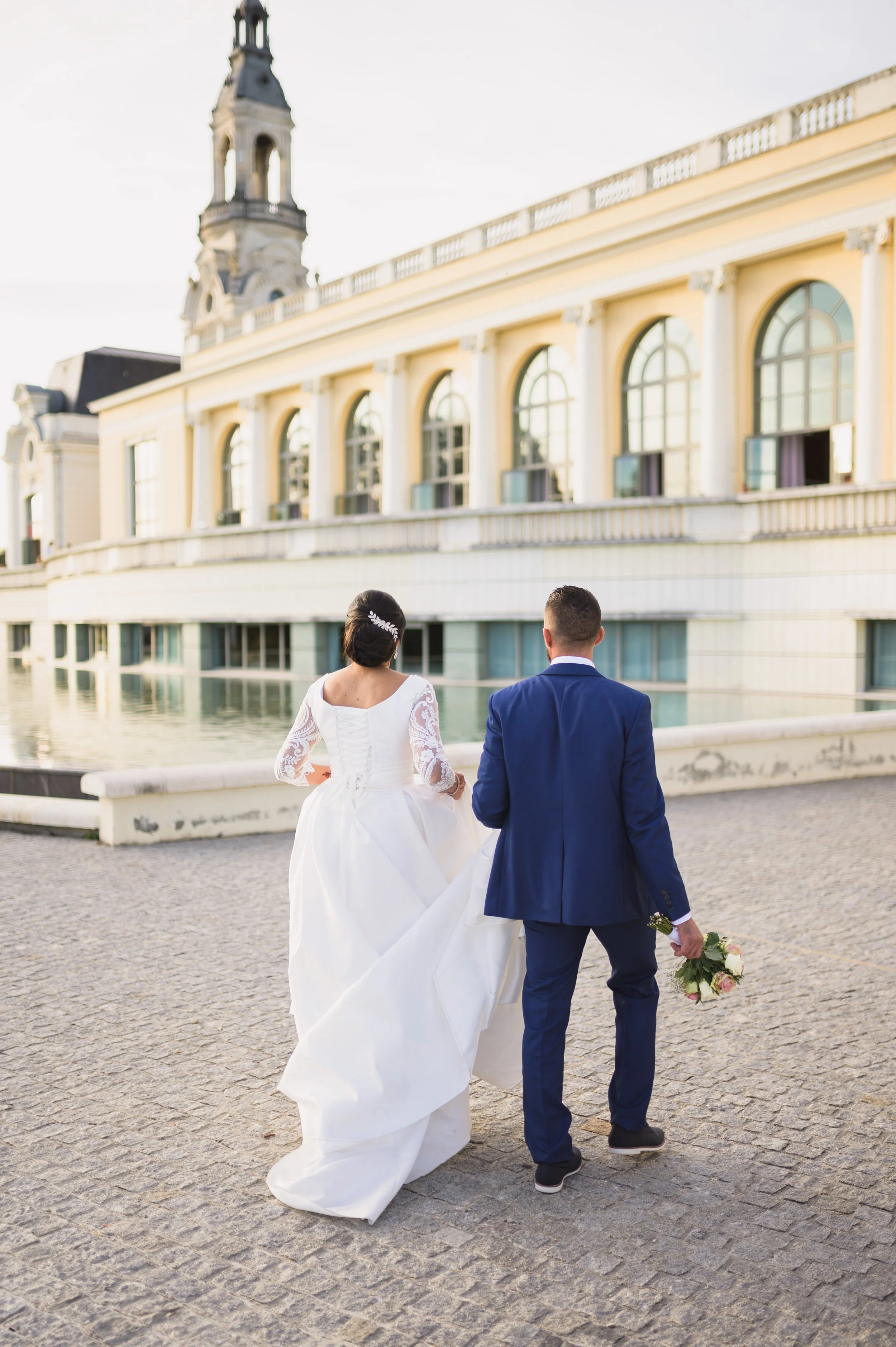 Bride in white gown and groom in blue suit walk together by water, holding bouquet, near historic building with large windows and clock tower.