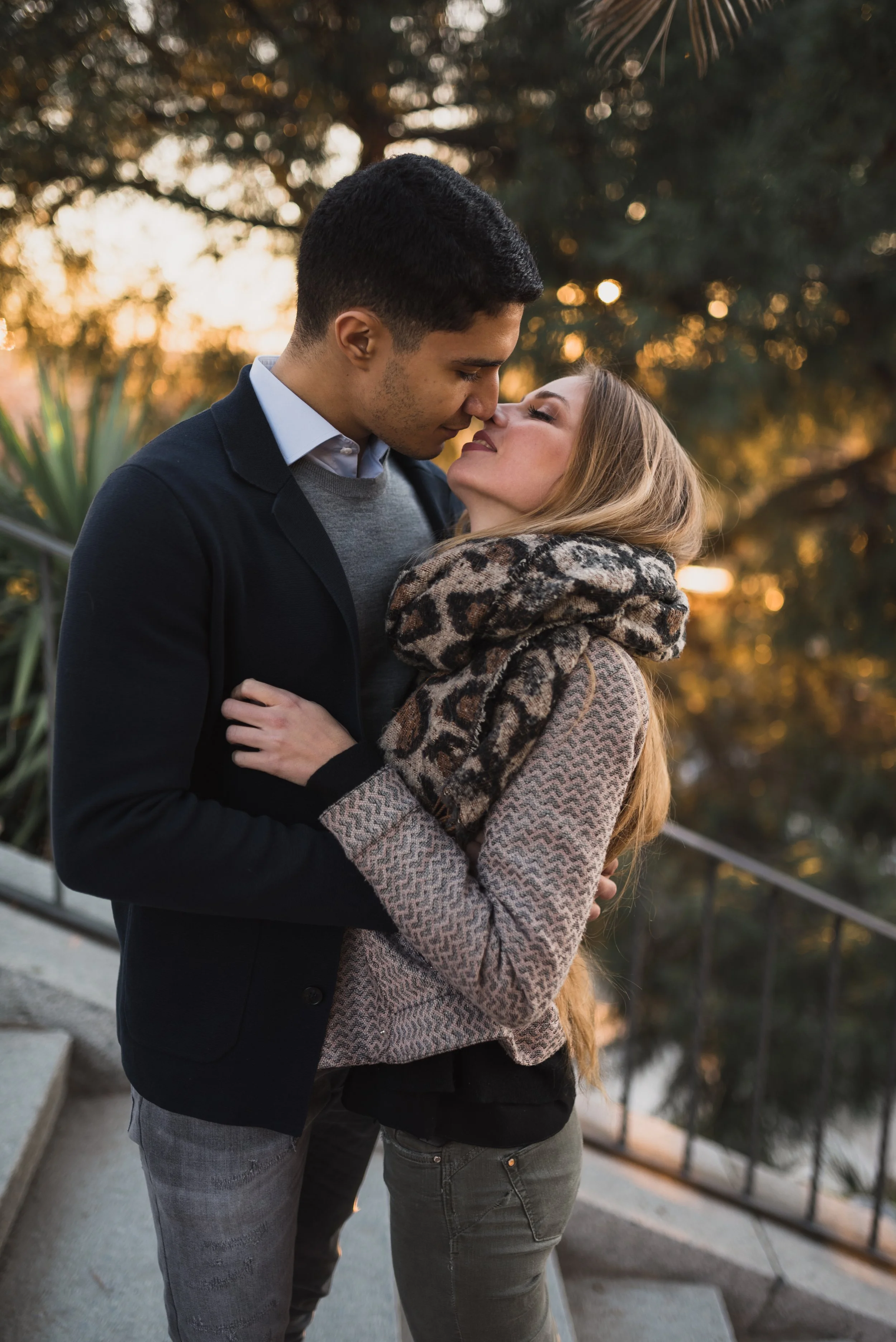 A young couple sharing an intimate moment outdoors at sunset, their faces close together, surrounded by trees.
