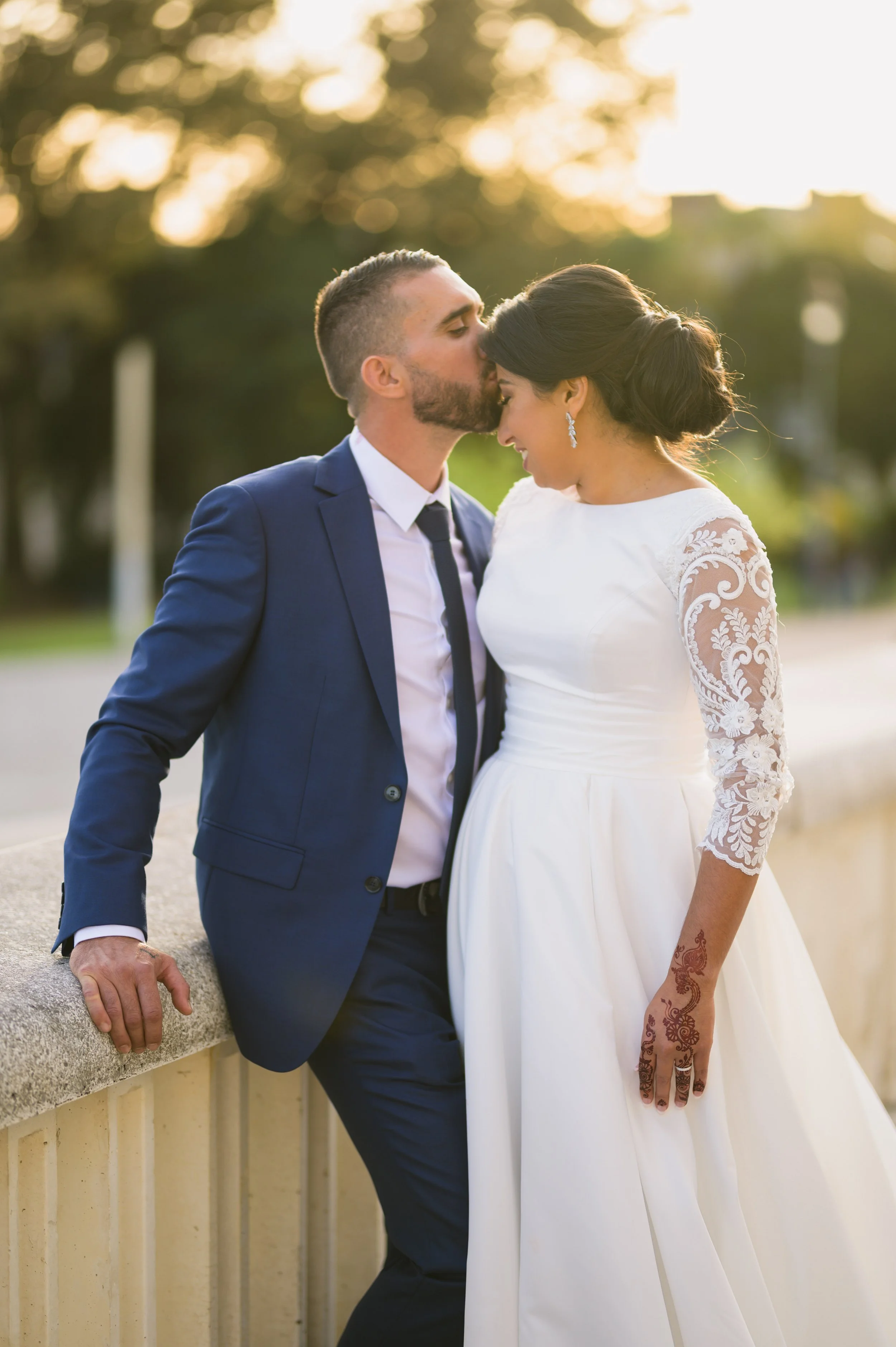 Couple on their wedding day, the groom in a blue suit kissing the bride on the forehead, she is wearing a white wedding dress with lace sleeves, standing outdoors during sunset.