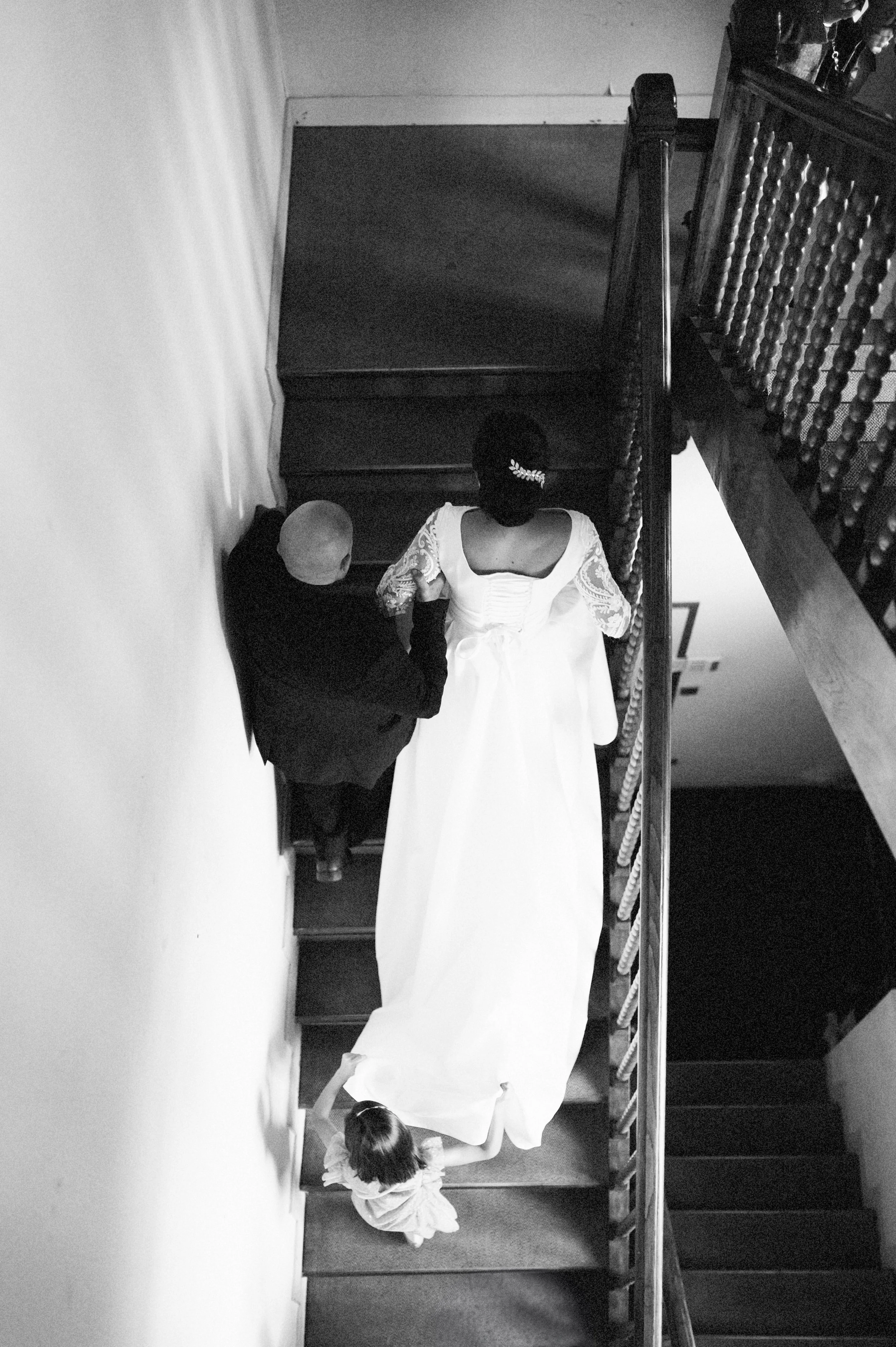 Black and white photo of a bride wearing a wedding dress with lace sleeves, walking up a staircase with two adults and a young girl accompanying her.