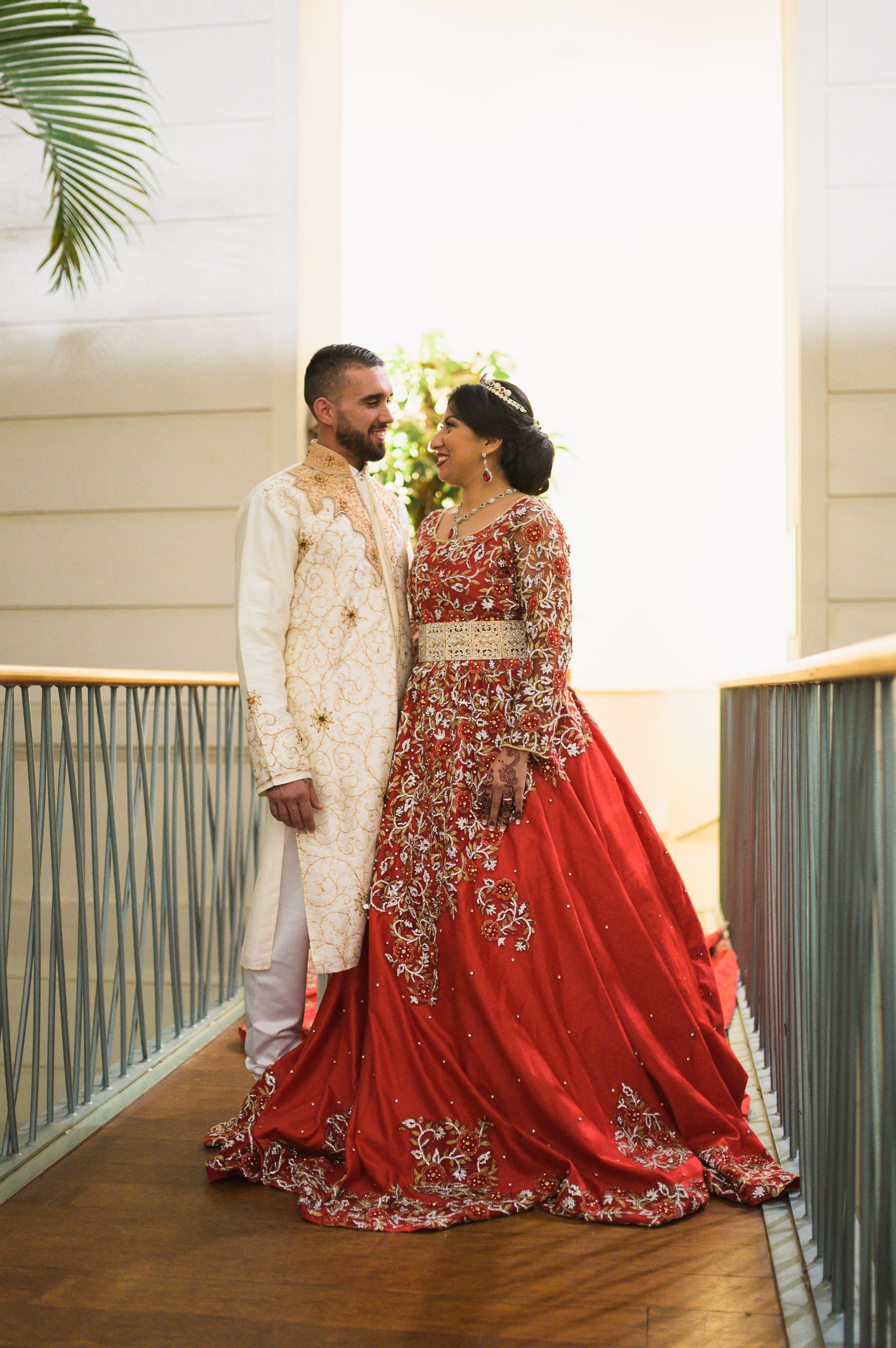 A couple dressed in traditional Indian wedding attire, standing close and smiling at each other, inside a bright, airy venue.