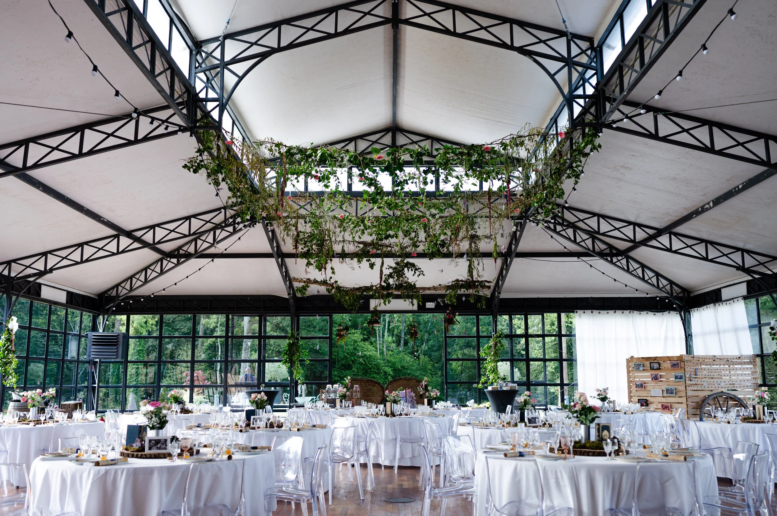 Indoor event space with white round tables covered in white tablecloths, decorated with floral centerpieces and table settings. Clear chairs surround the tables. The ceiling has black metal frameworks and is adorned with hanging greenery and string l
