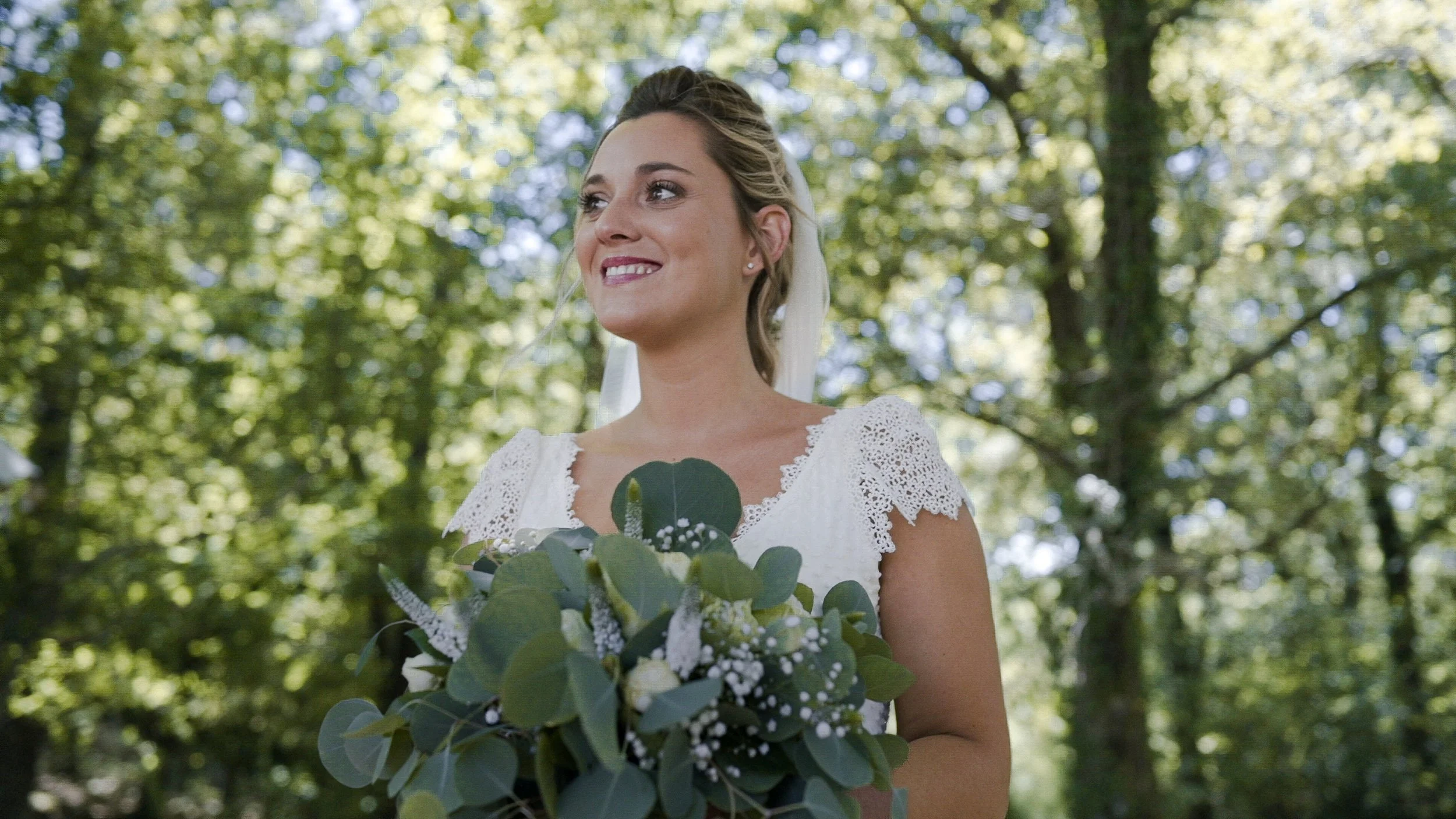 A smiling woman in a white lace wedding dress holding a bouquet of green leaves and white flowers outdoors with trees in the background.