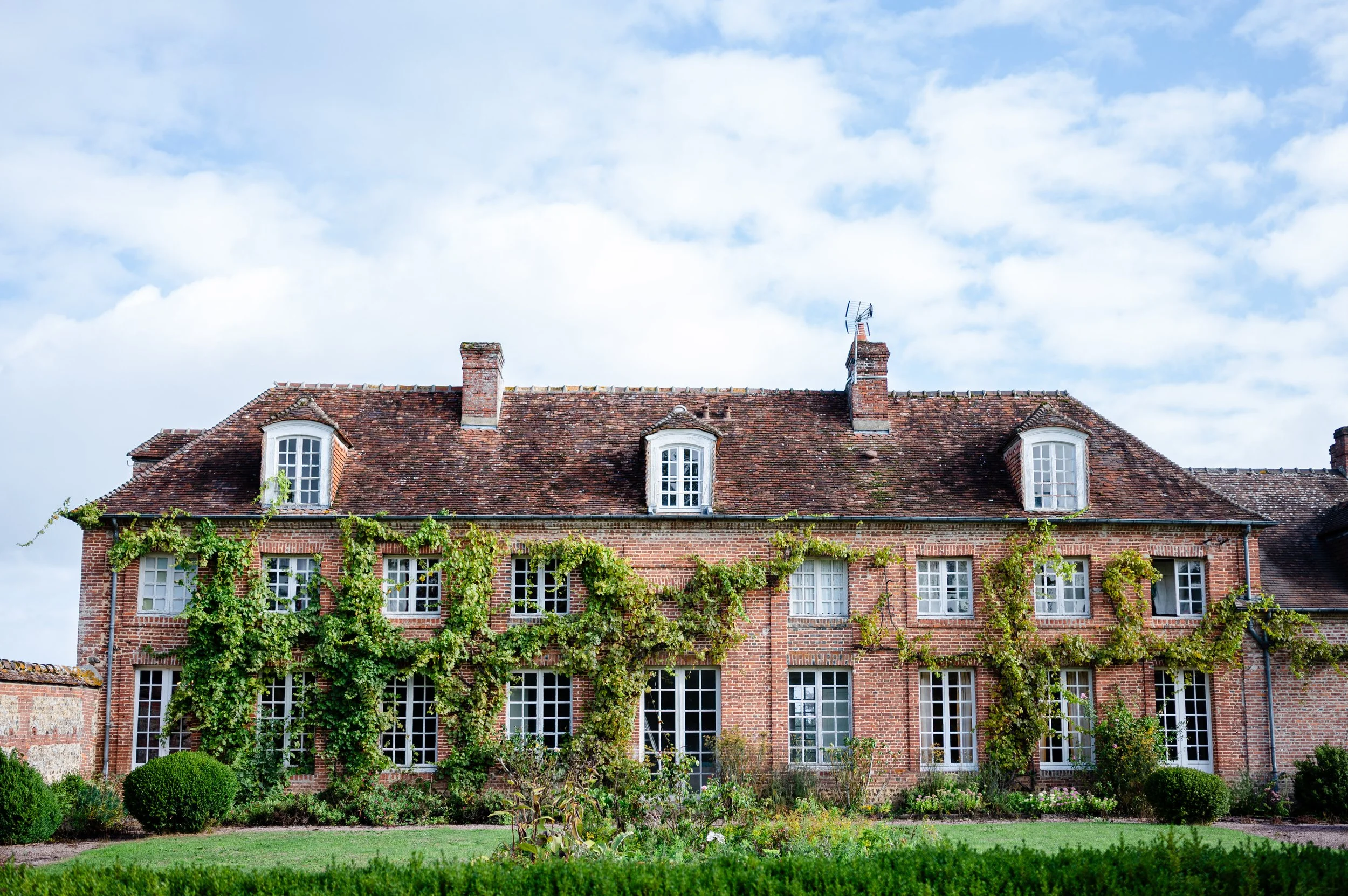 A large brick house with climbing vines growing along the front facade, multiple windows, and a red tiled roof under a partly cloudy sky.