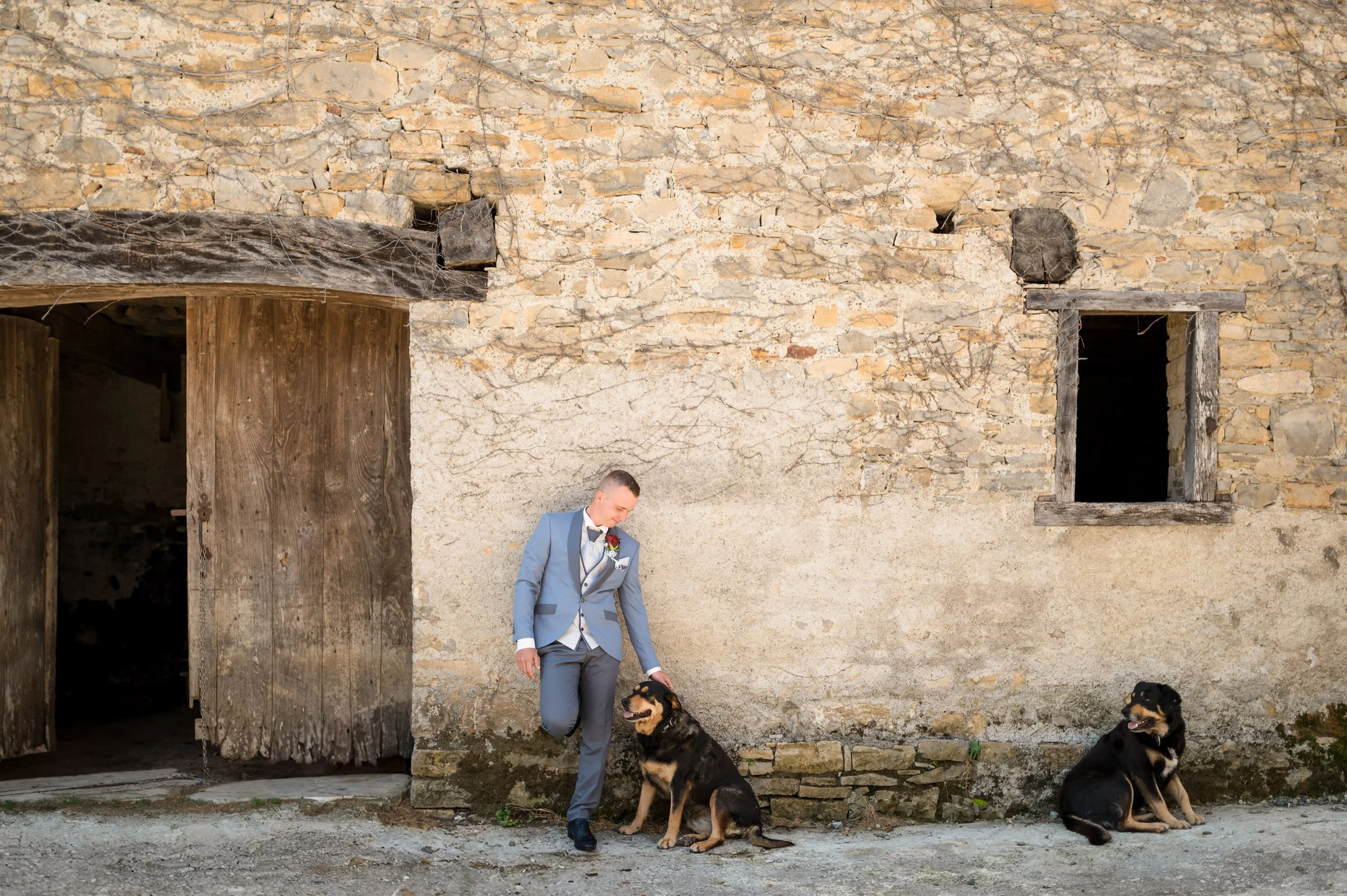 A man in a gray suit standing next to a large black and tan dog against an old stone wall.