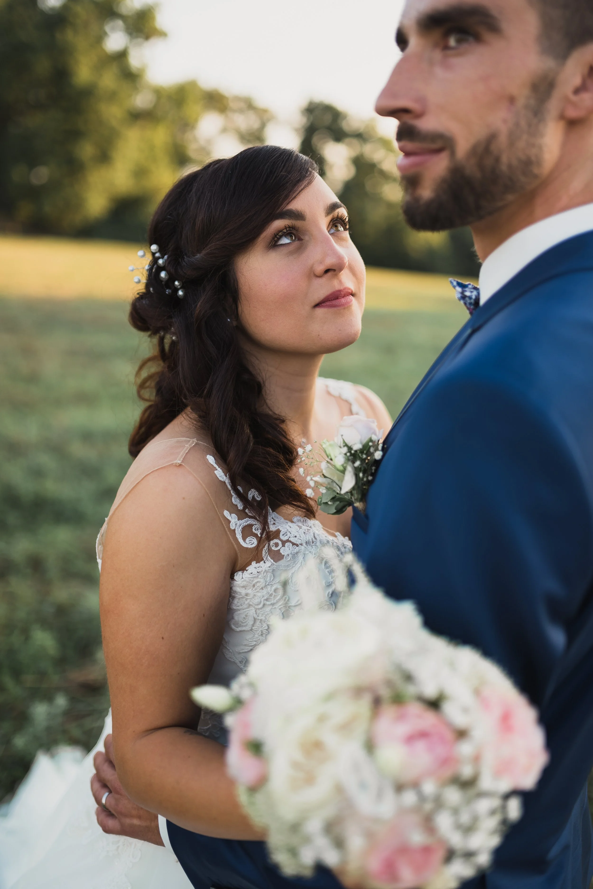 Bride and groom on wedding day outdoors, bride holding a bouquet, bride looking lovingly at groom, trees and sunlight in background.