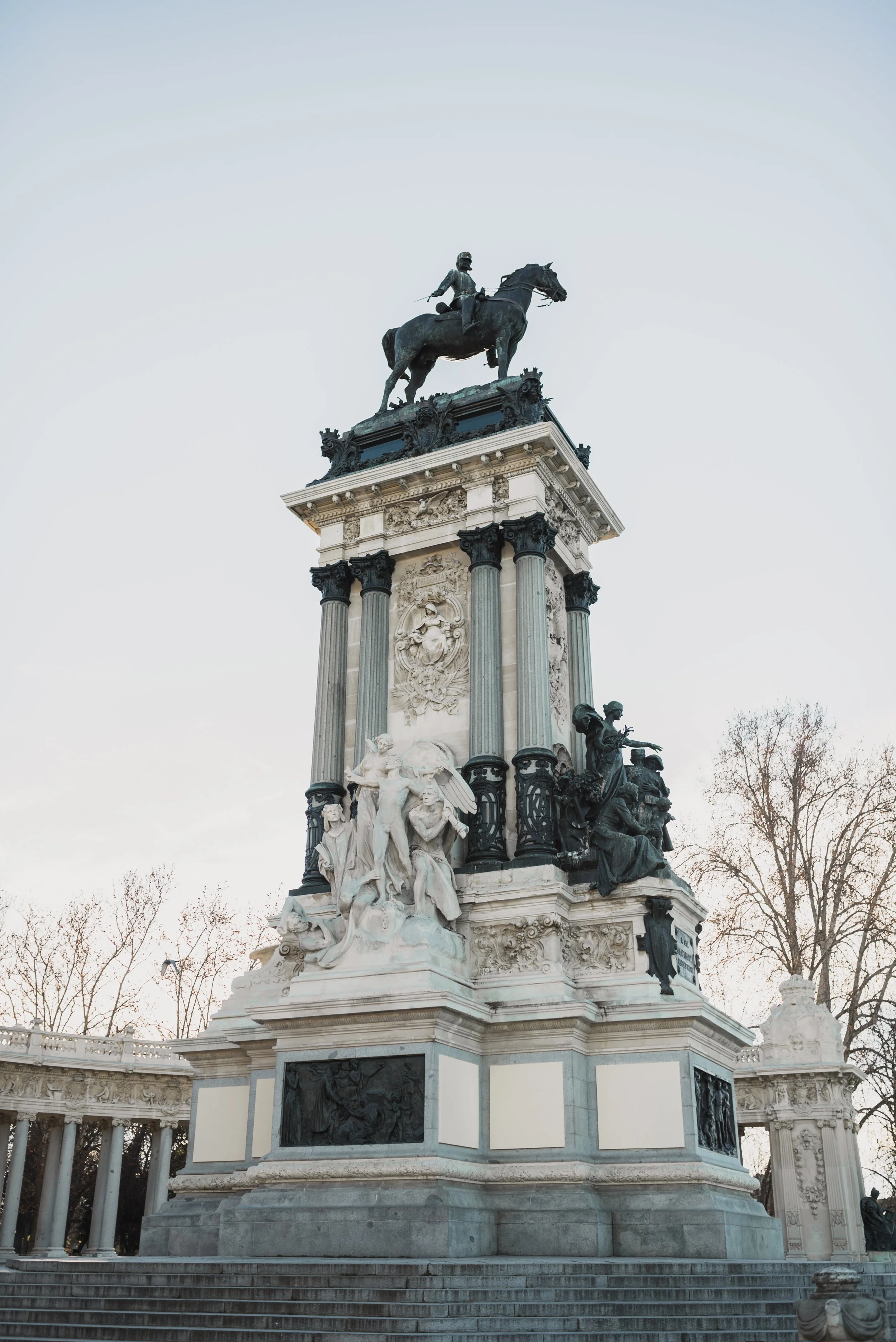 A tall monument with a statue of a man on a horse at the top, surrounded by ornate sculptures and columns, set against a light sky with bare trees nearby.
