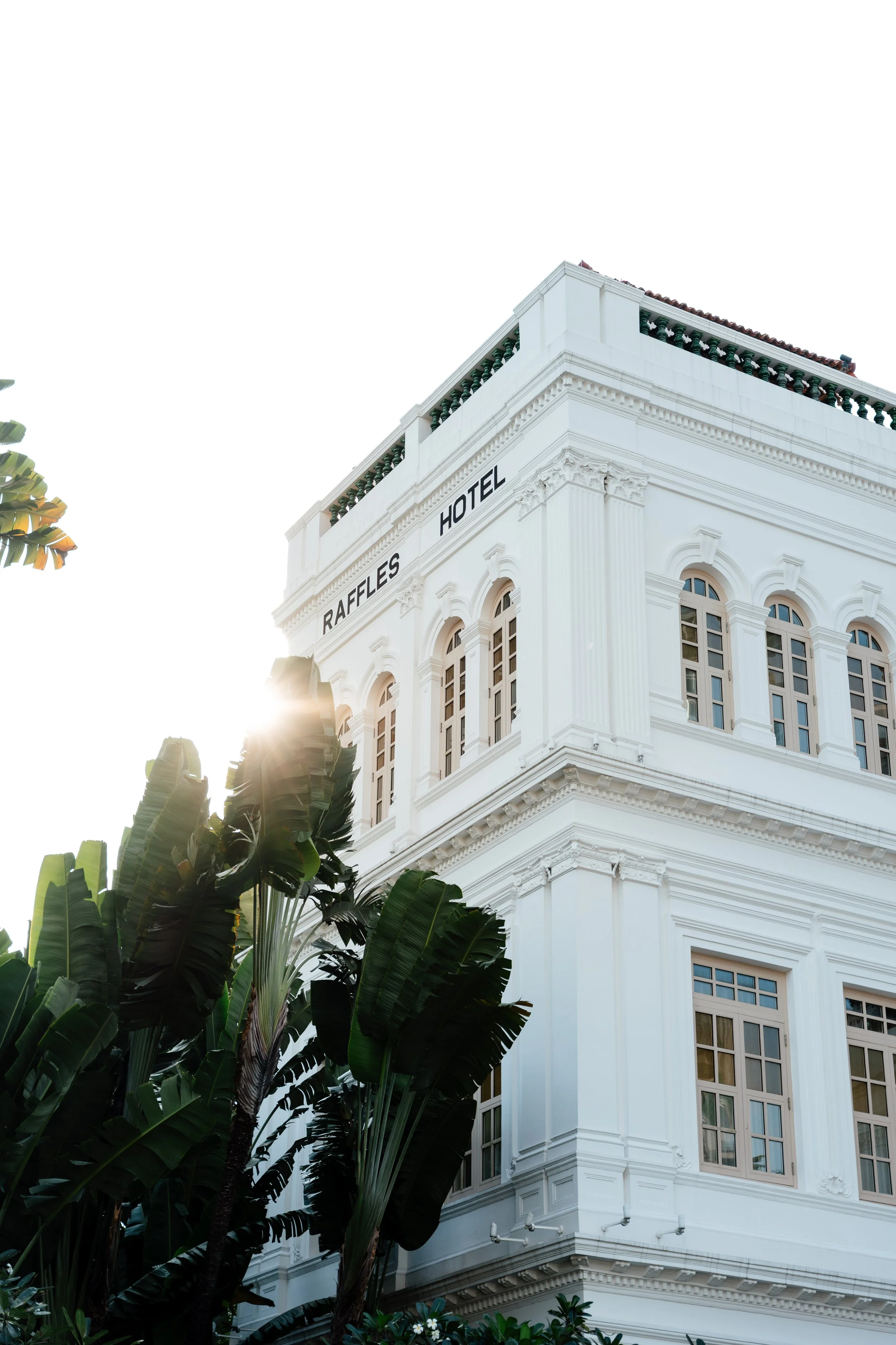White historic building with the sign "Raffles Hotel" on the corner, surrounded by greenery, with sunlight peeking through trees in the background.