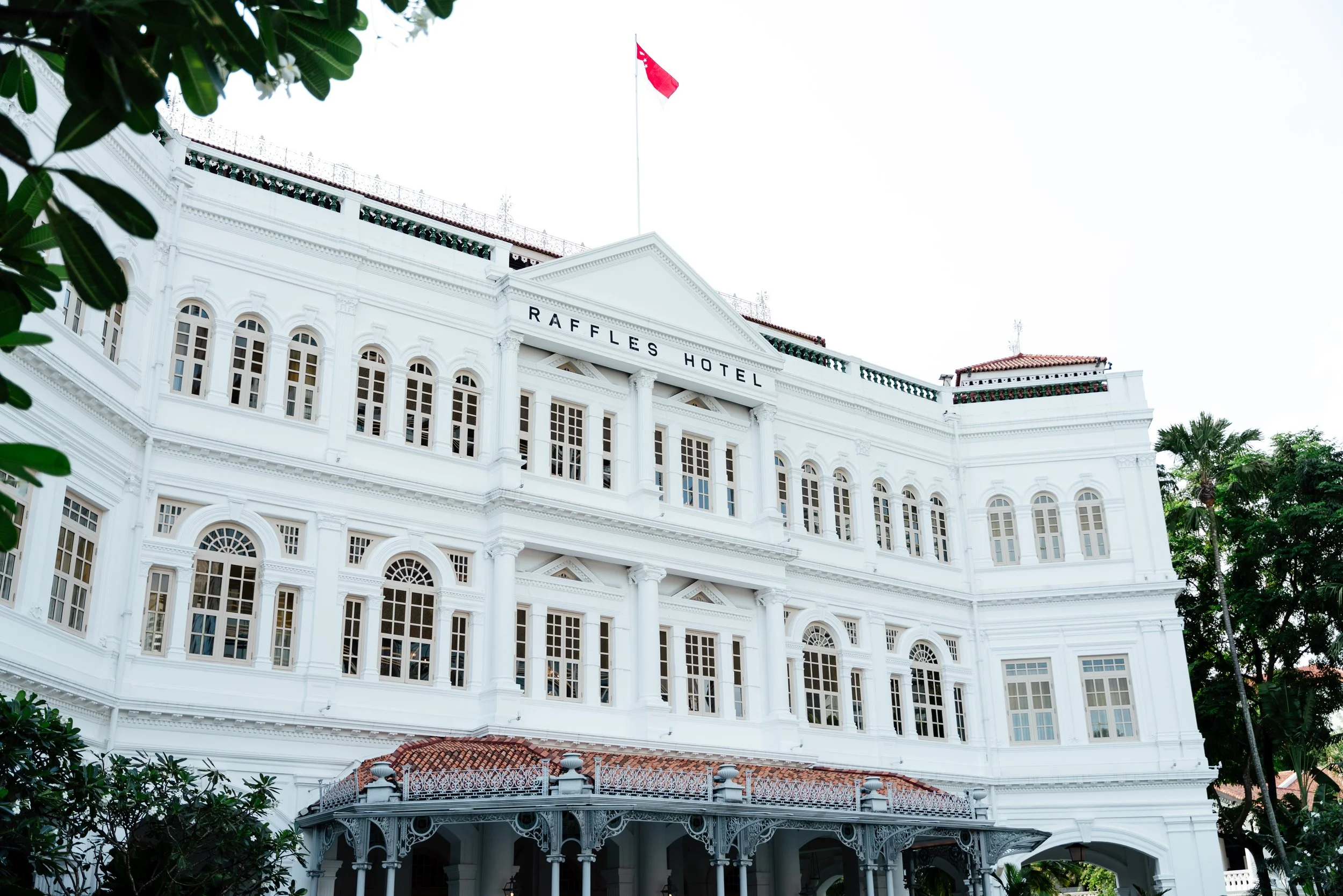White, historic building labeled Raffles Hotel with colonial architecture, numerous tall windows, a red-tiled roof, surrounded by greenery.