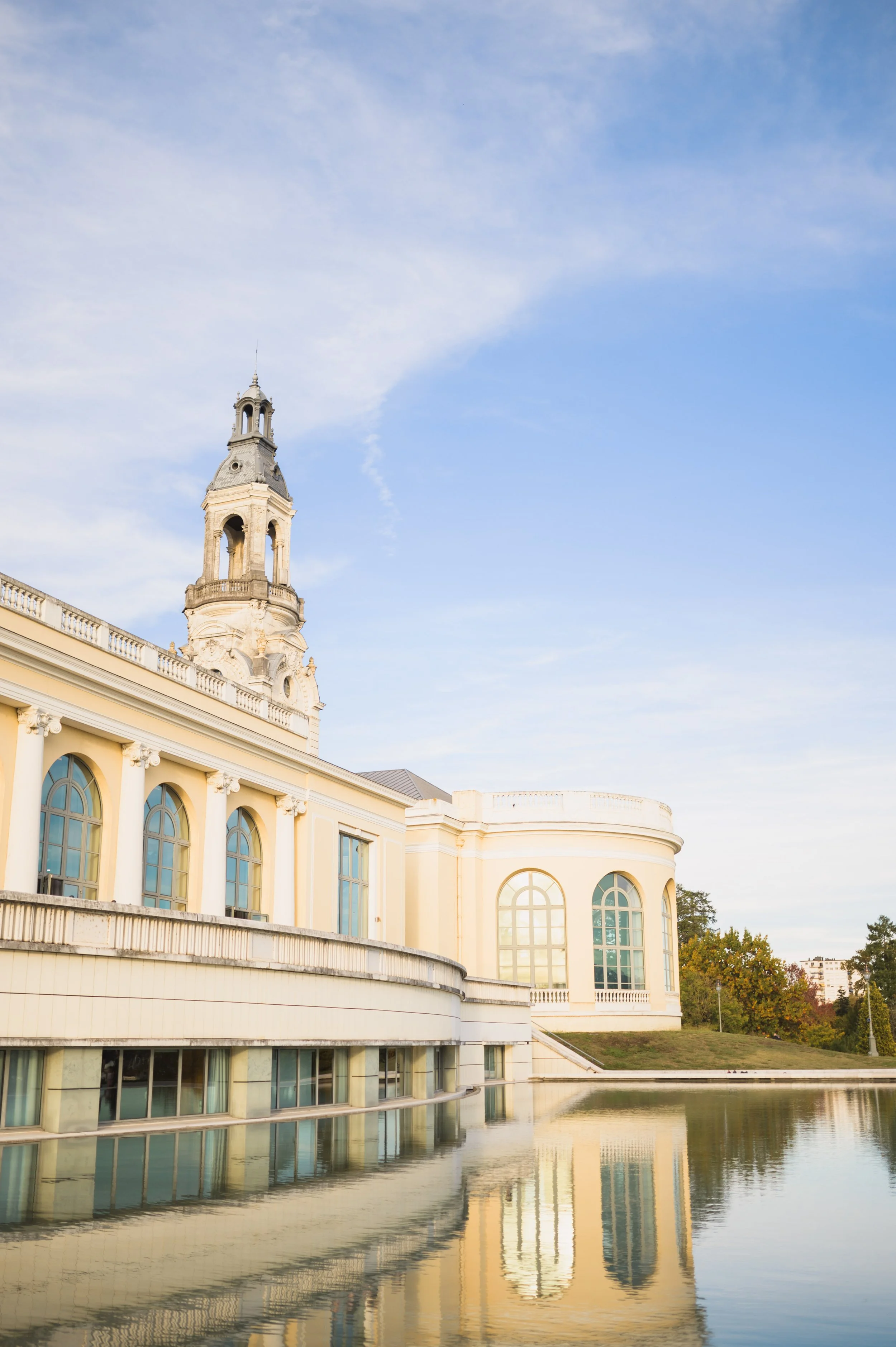 A classic European-style building with a tall bell tower, arched windows, cream-colored walls, and a water reflection, set against a partly cloudy sky.