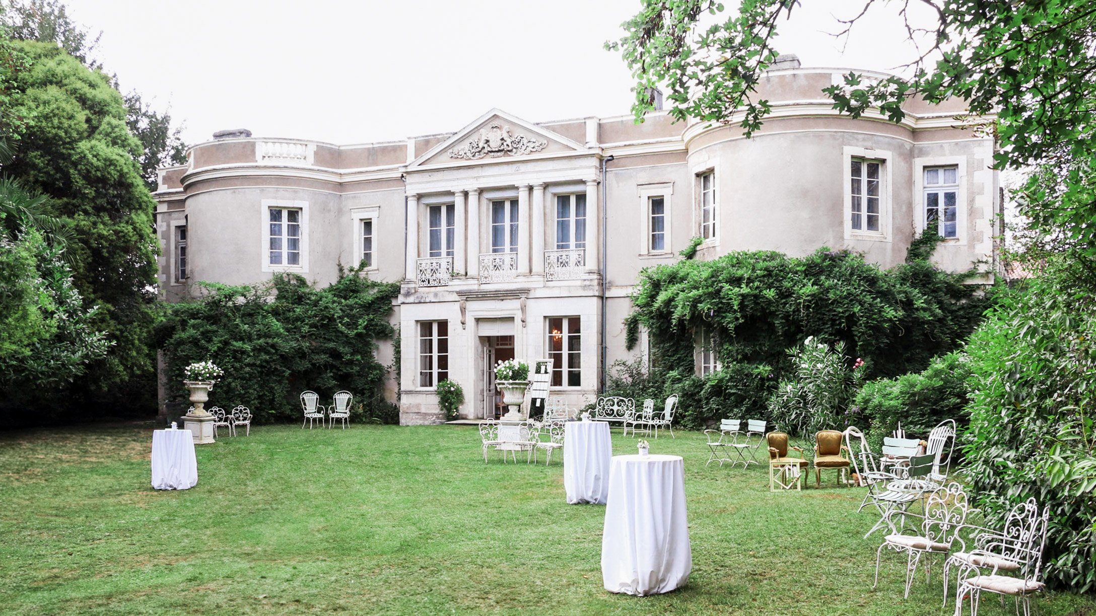Elegant garden with white tables, chairs, and planters in front of a historic mansion surrounded by lush greenery.