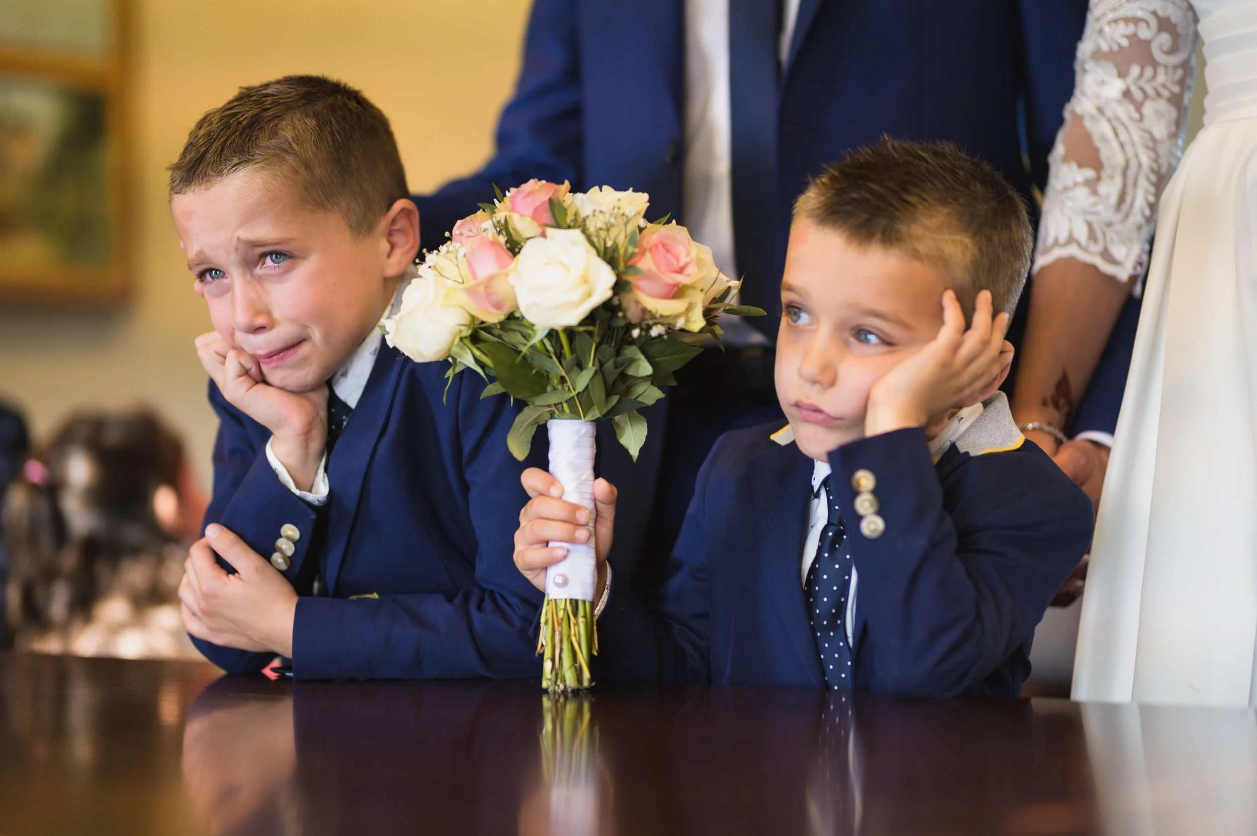 Two young boys in navy suits sat at a table during a wedding or special event, with one holding a bouquet of pink and white roses, both appearing bored or upset.