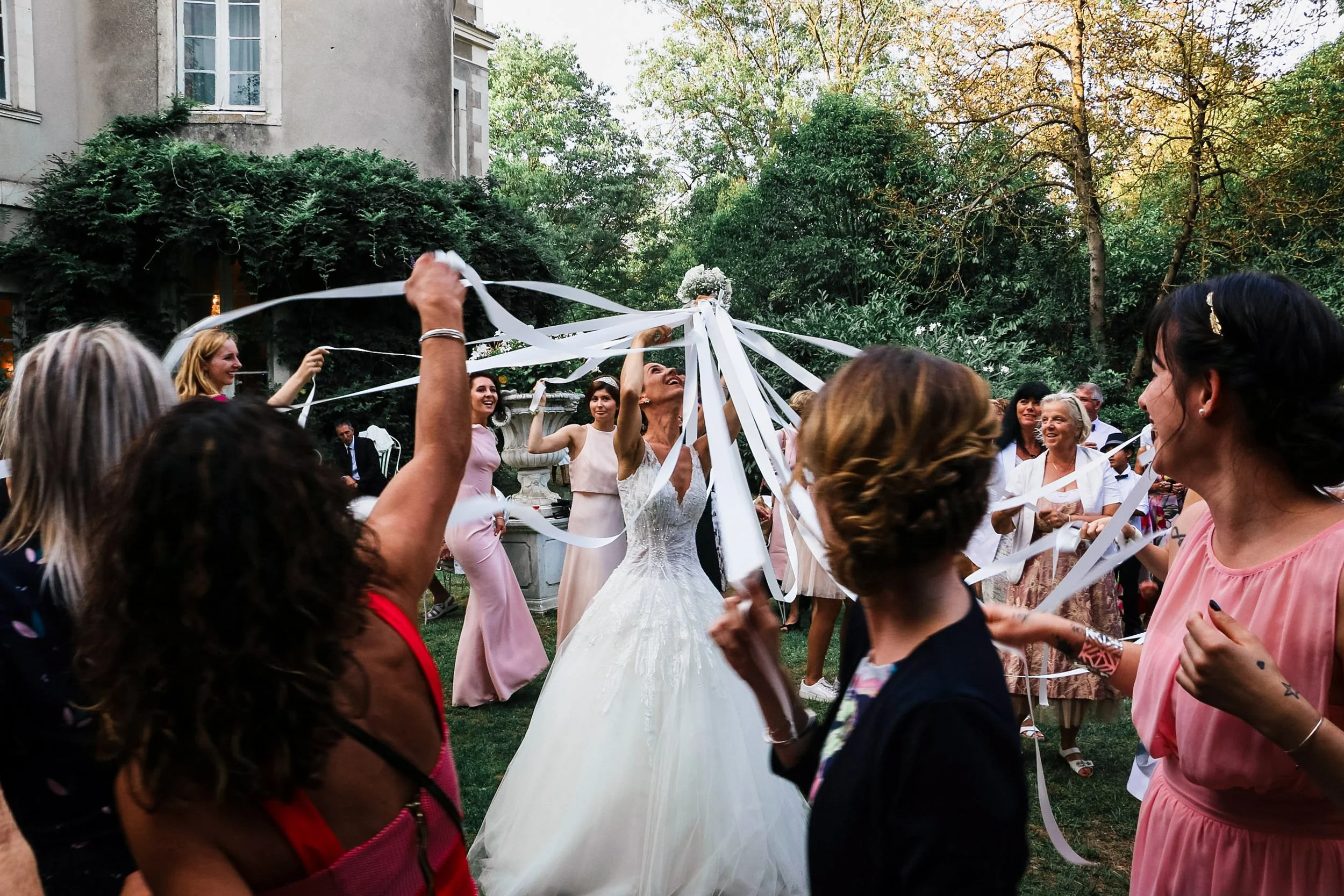 Bride and wedding guests celebrating with ribbons outdoors in a garden, trees in background.