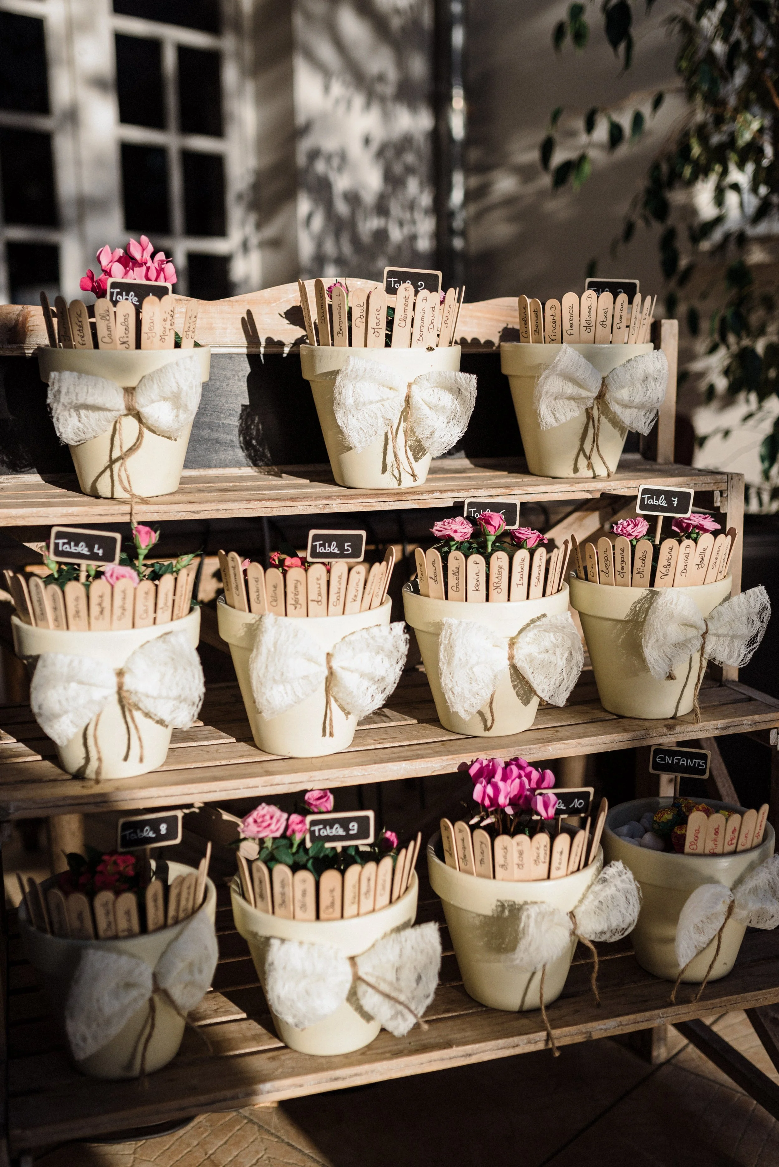 Decorative flower pots with wooden plant markers, arranged on wooden shelves, with small chalkboard signs indicating table numbers and pink flowers inside some pots.