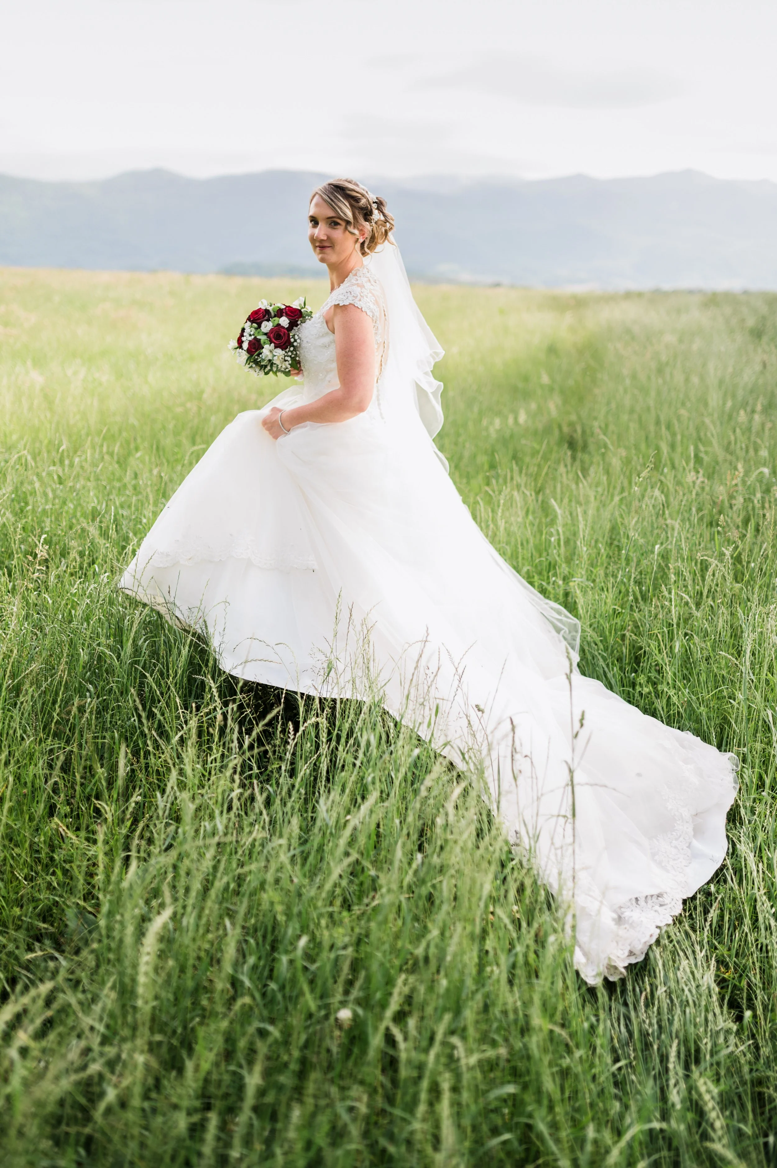 A bride in a white wedding gown holding a bouquet of red and white flowers, standing in a green field with mountains in the background.