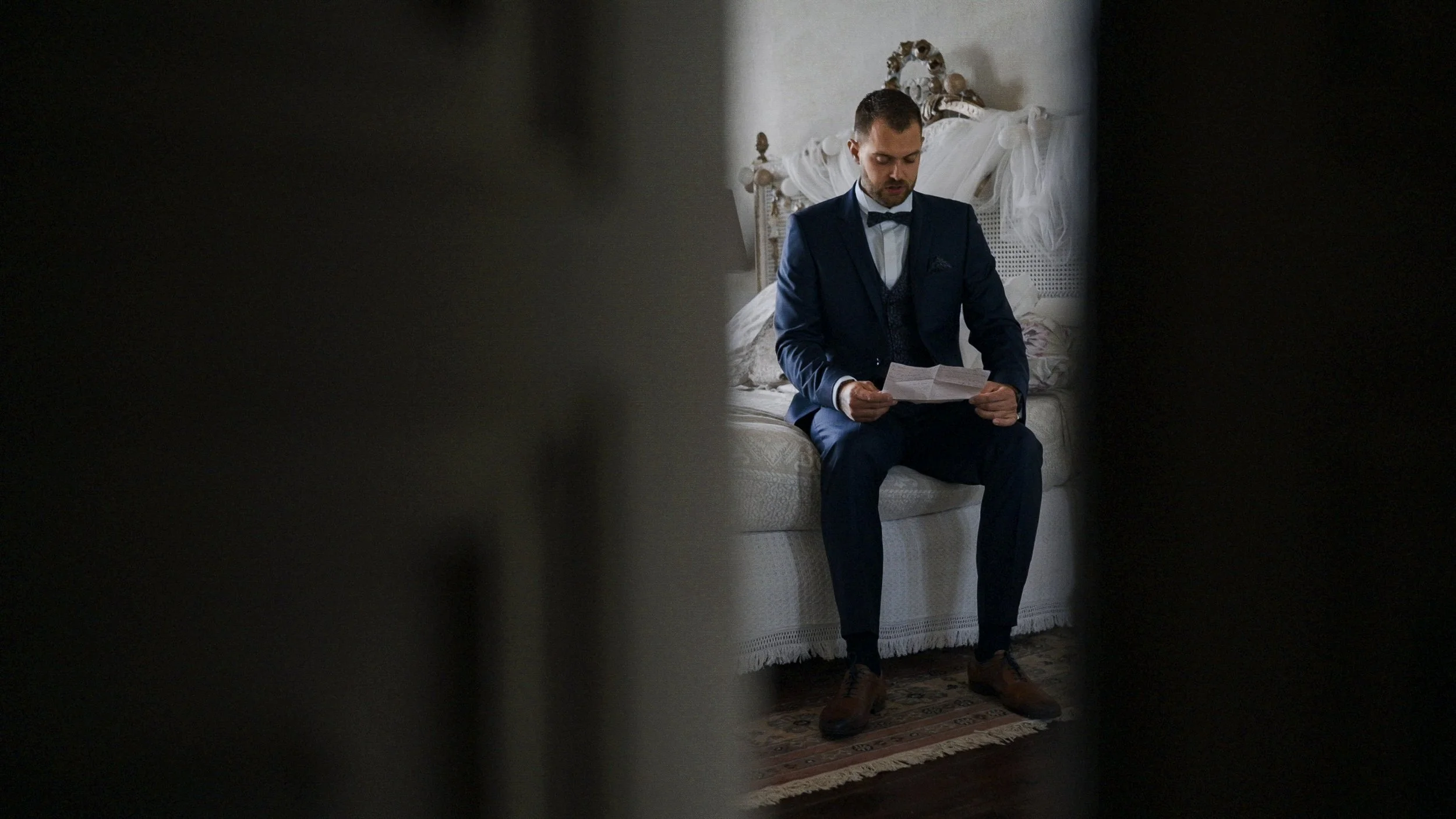 A man in a dark blue suit sitting on a bed, reading a paper, viewed through a partially opened door.