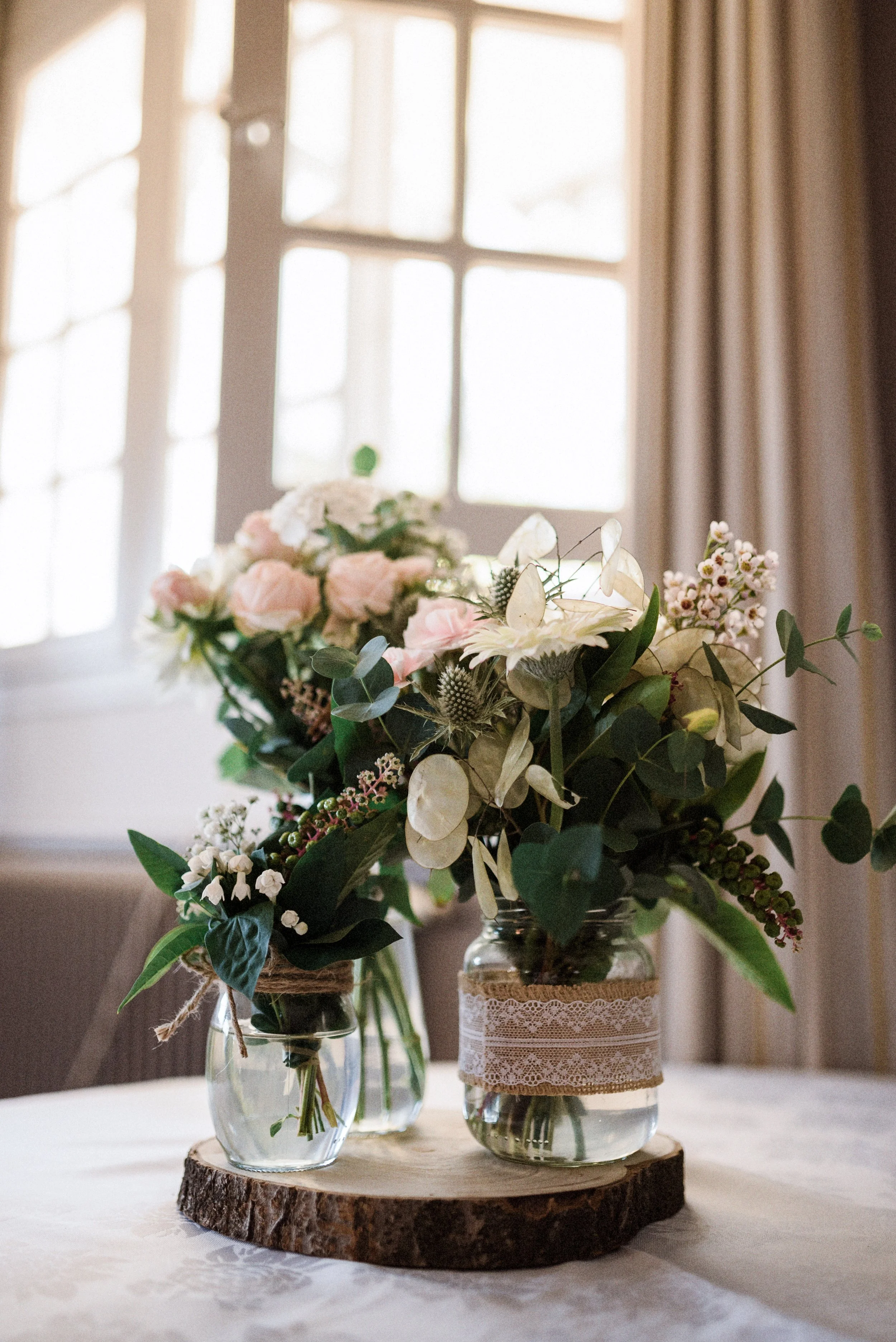 Two glass jars with floral arrangements on a wooden slab, with a background of a window and curtains.
