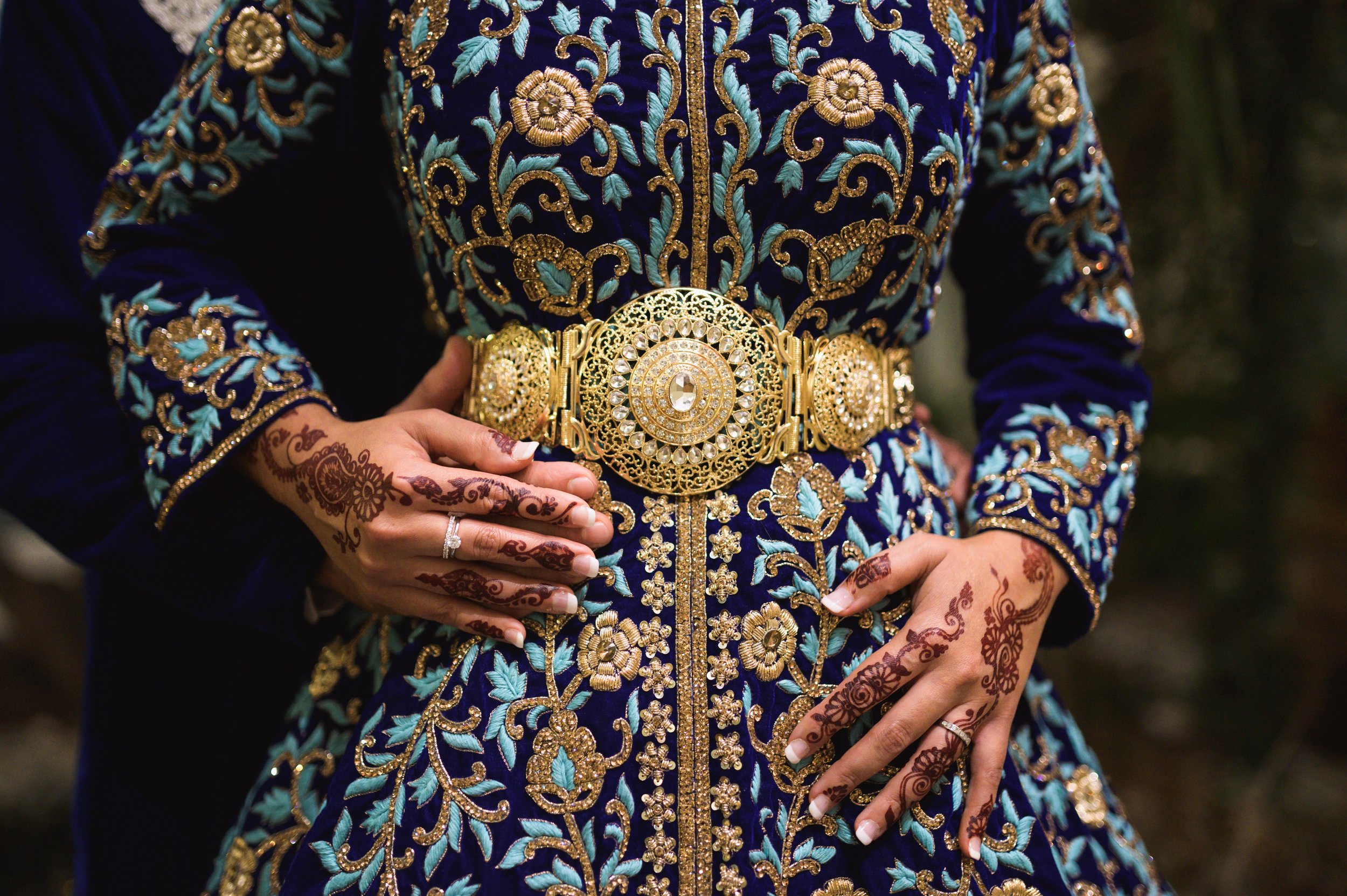 Close-up of a woman wearing a richly embroidered blue velvet dress with gold floral embroidery, accessorized with a large, ornate gold belt with embedded stones, and decorated with henna designs on her hands.