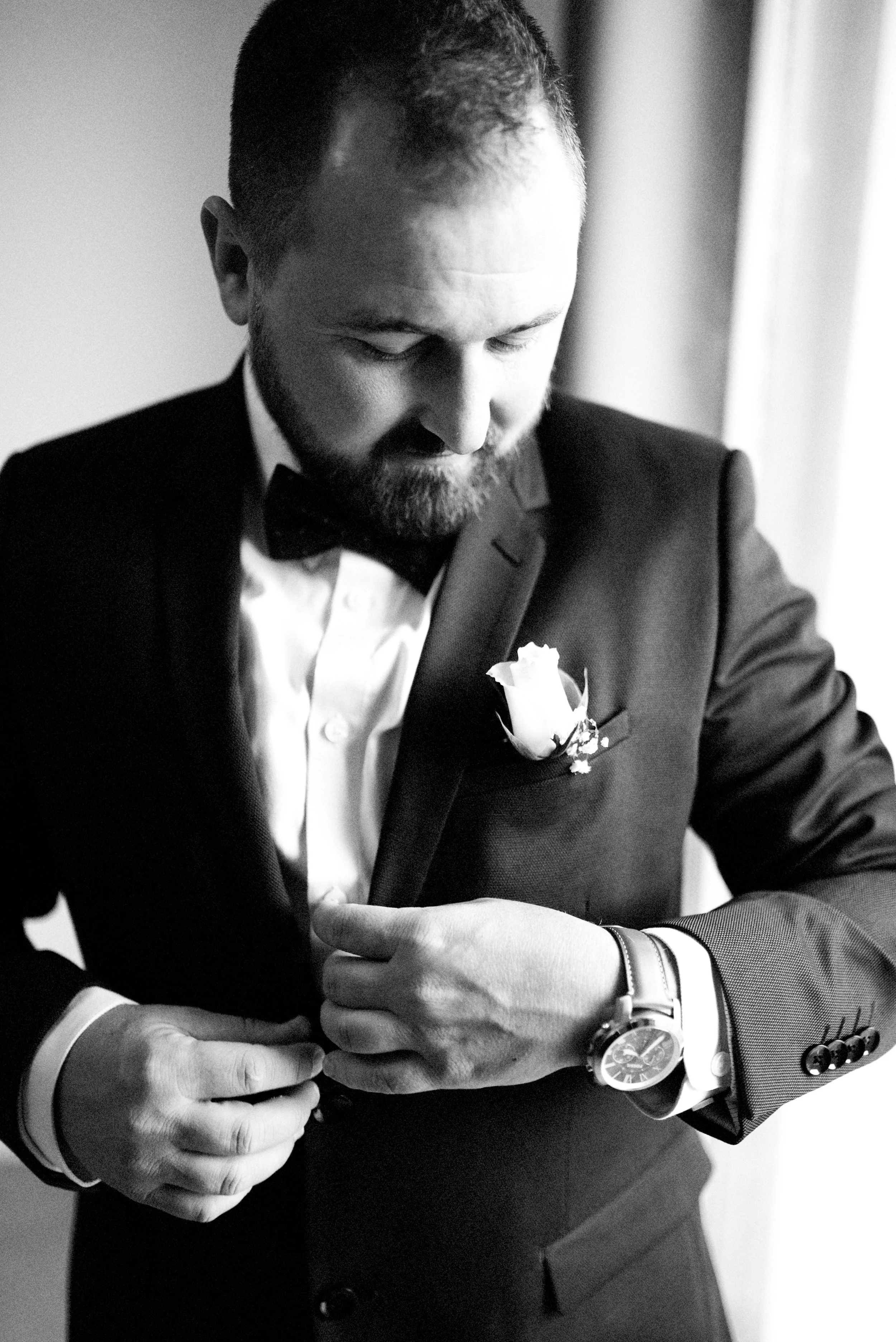 A man in a tuxedo with a boutonniere, adjusting his sleeve, looking down thoughtfully.