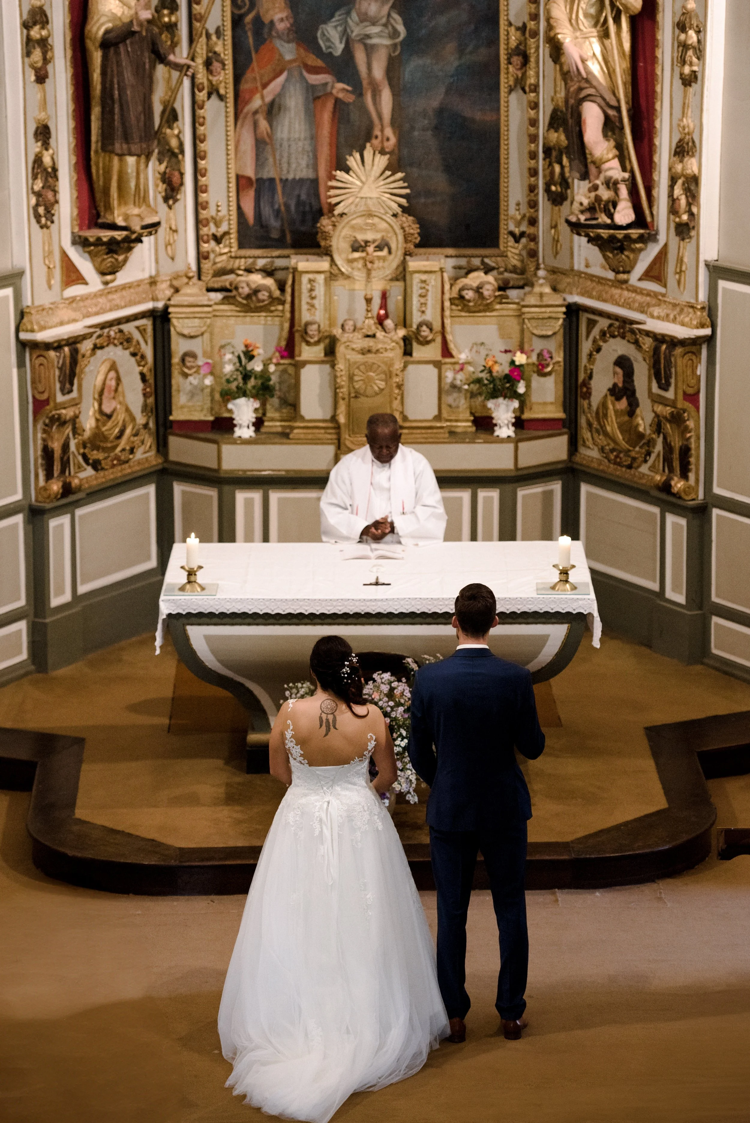 A bride and groom standing before a priest during a wedding ceremony inside a church. The bride is wearing a white wedding gown with lace details, and the groom is in a dark suit. They are facing the priest who is standing behind an altar decorated w