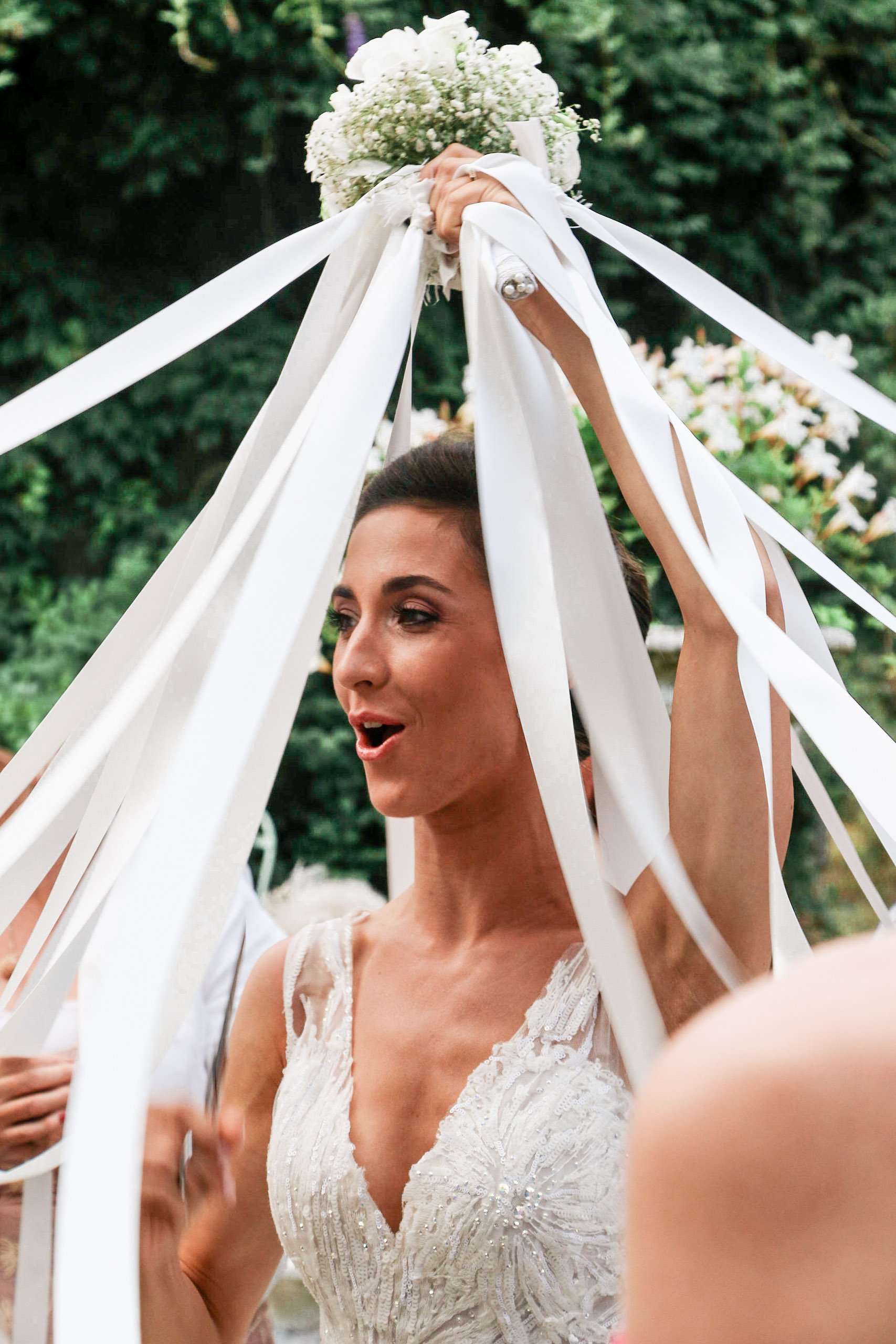 A woman in a wedding dress holding a bouquet of flowers above her head during a celebration outdoors.