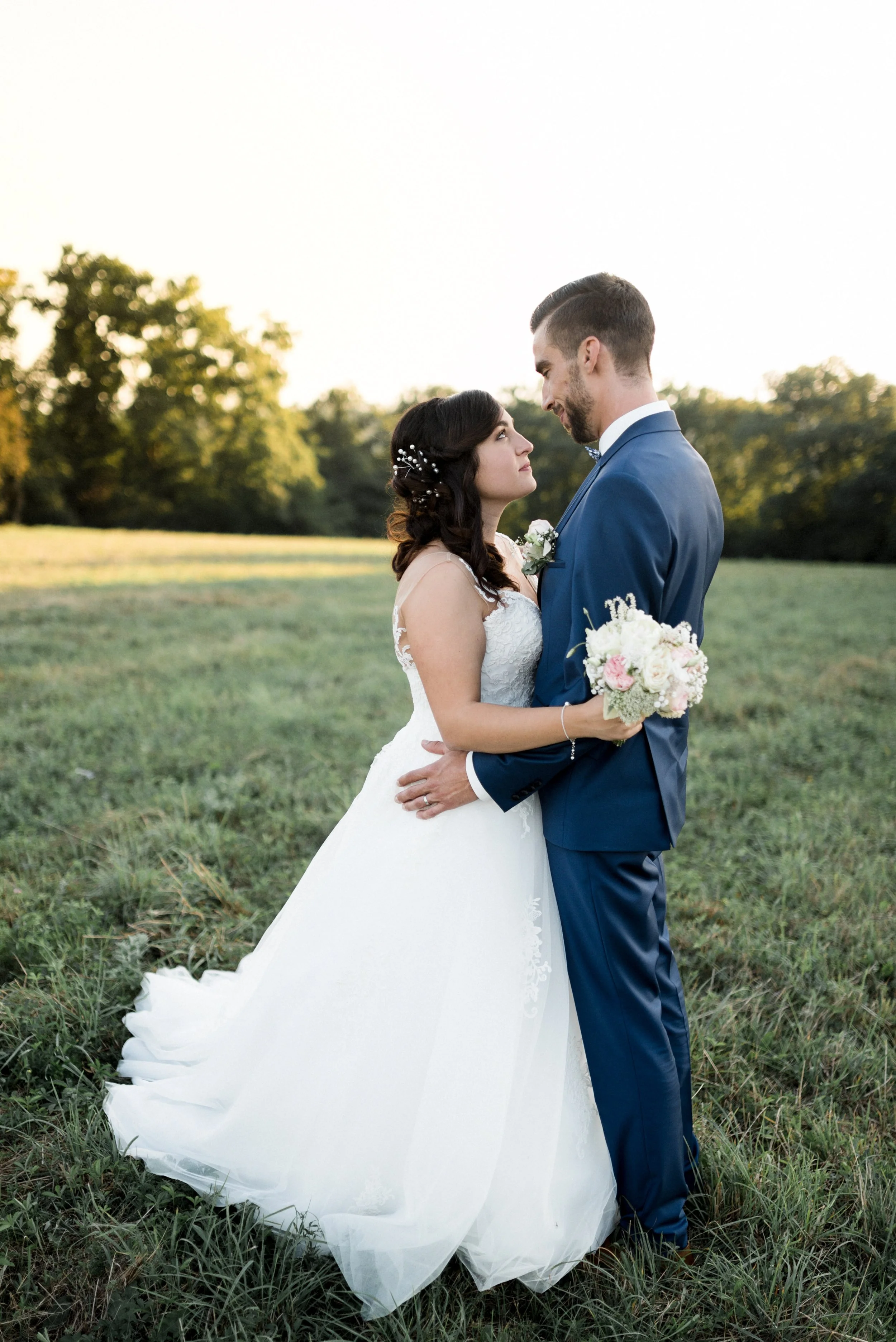 A bride and groom embrace outdoors at sunset, with the bride holding a bouquet of white and pink flowers and wearing a white wedding gown, and the groom in a blue suit. They gaze into each other's eyes on a grassy field with trees in the background.