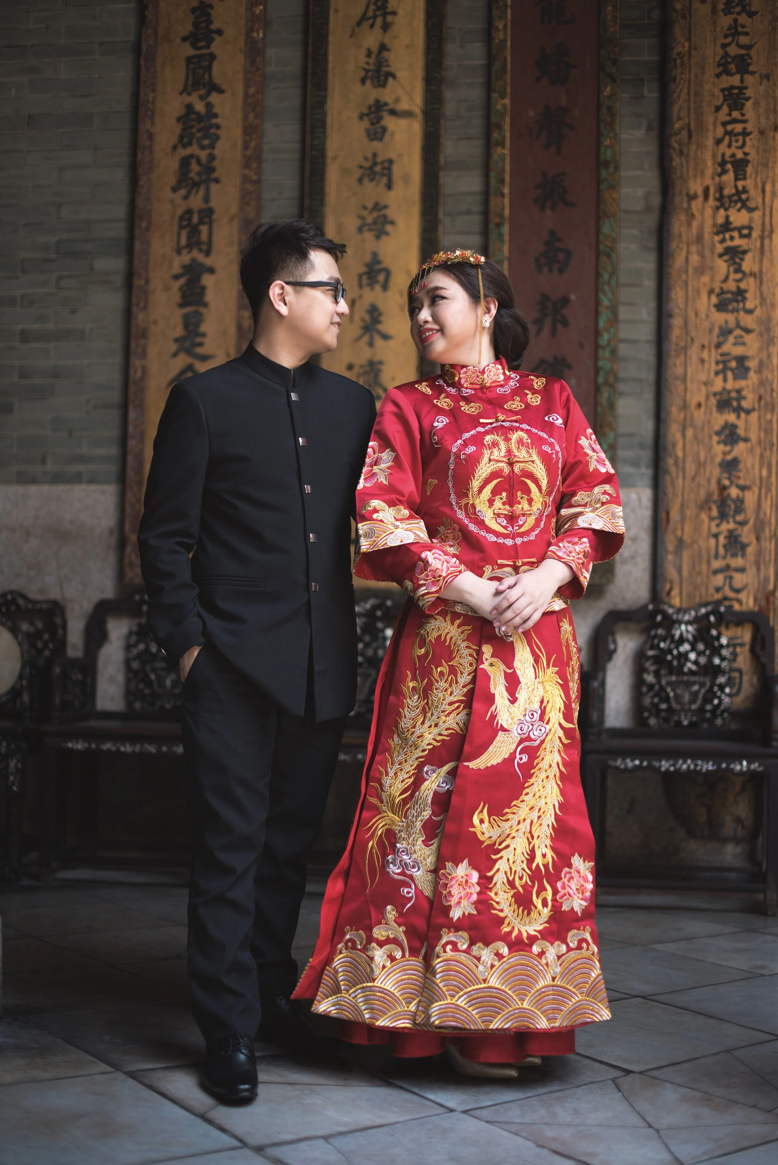 A couple dressed in traditional and modern Chinese wedding attire stands close together and looks at each other, inside a building with Chinese calligraphy banners on the wall behind them.