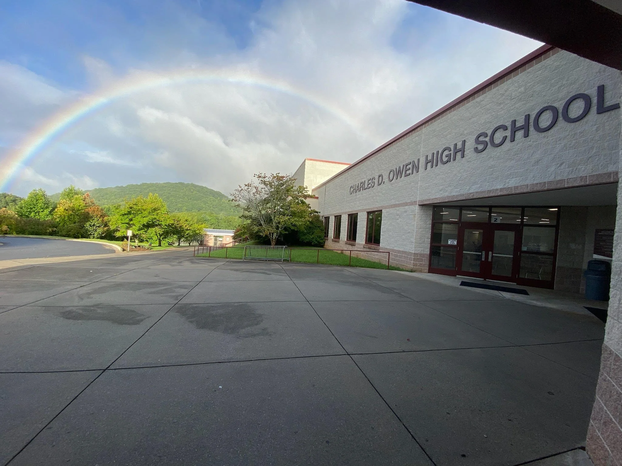 A rainbow arches over Charles D. Owen High School building with a mountain and trees in the background after rain. The school exterior is made of light-colored bricks with large windows and a glass door entrance.