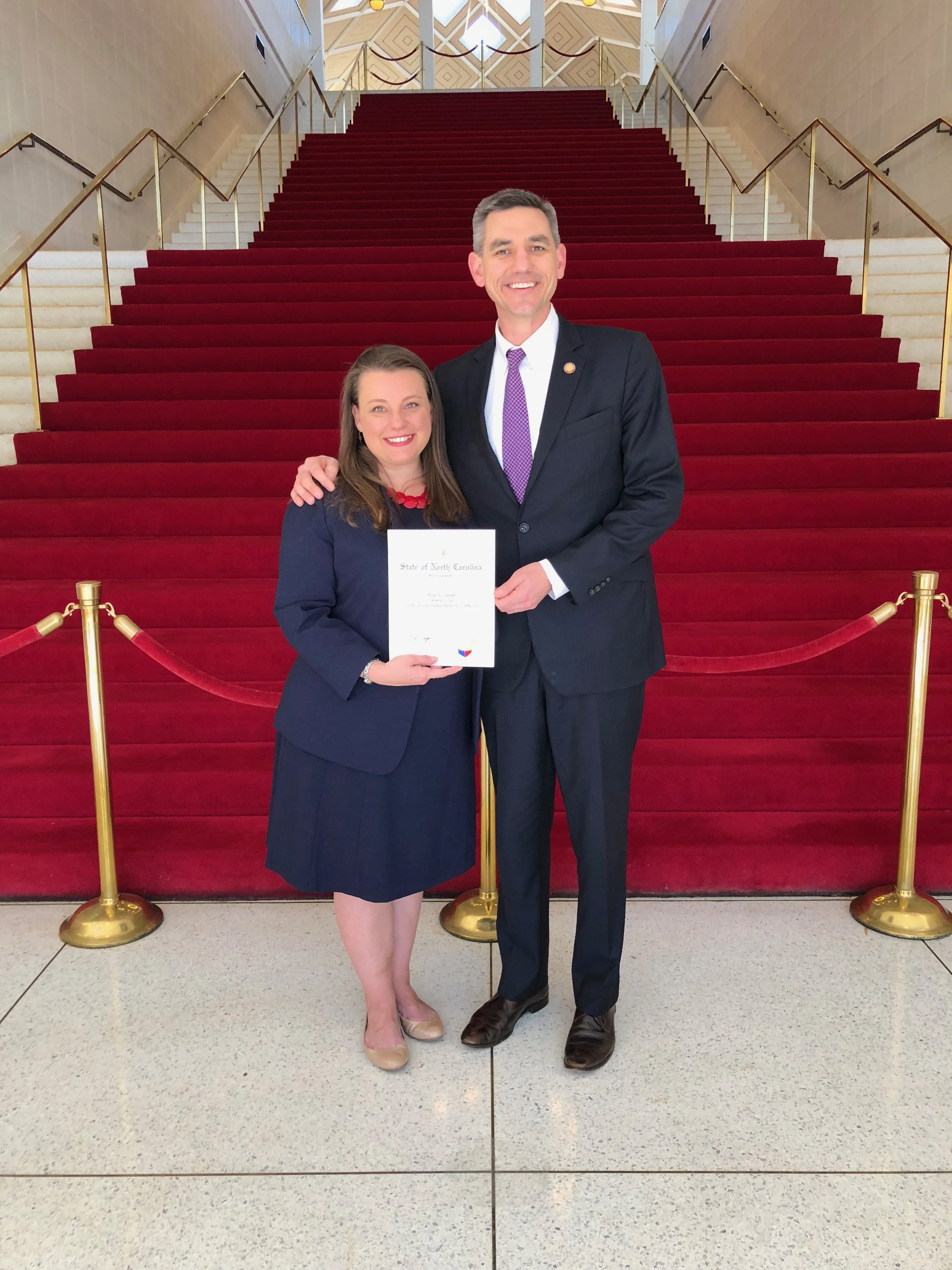 A woman and a man standing together on a red carpeted staircase in an official setting, with the woman holding a certificate. Both are smiling and dressed in formal attire.