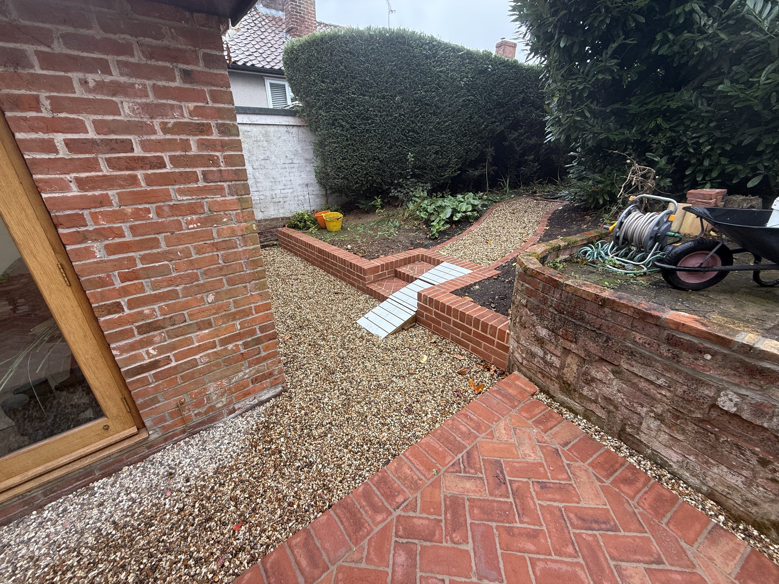 View of a backyard with brick and gravel pathways, greenery, and gardening tools.
