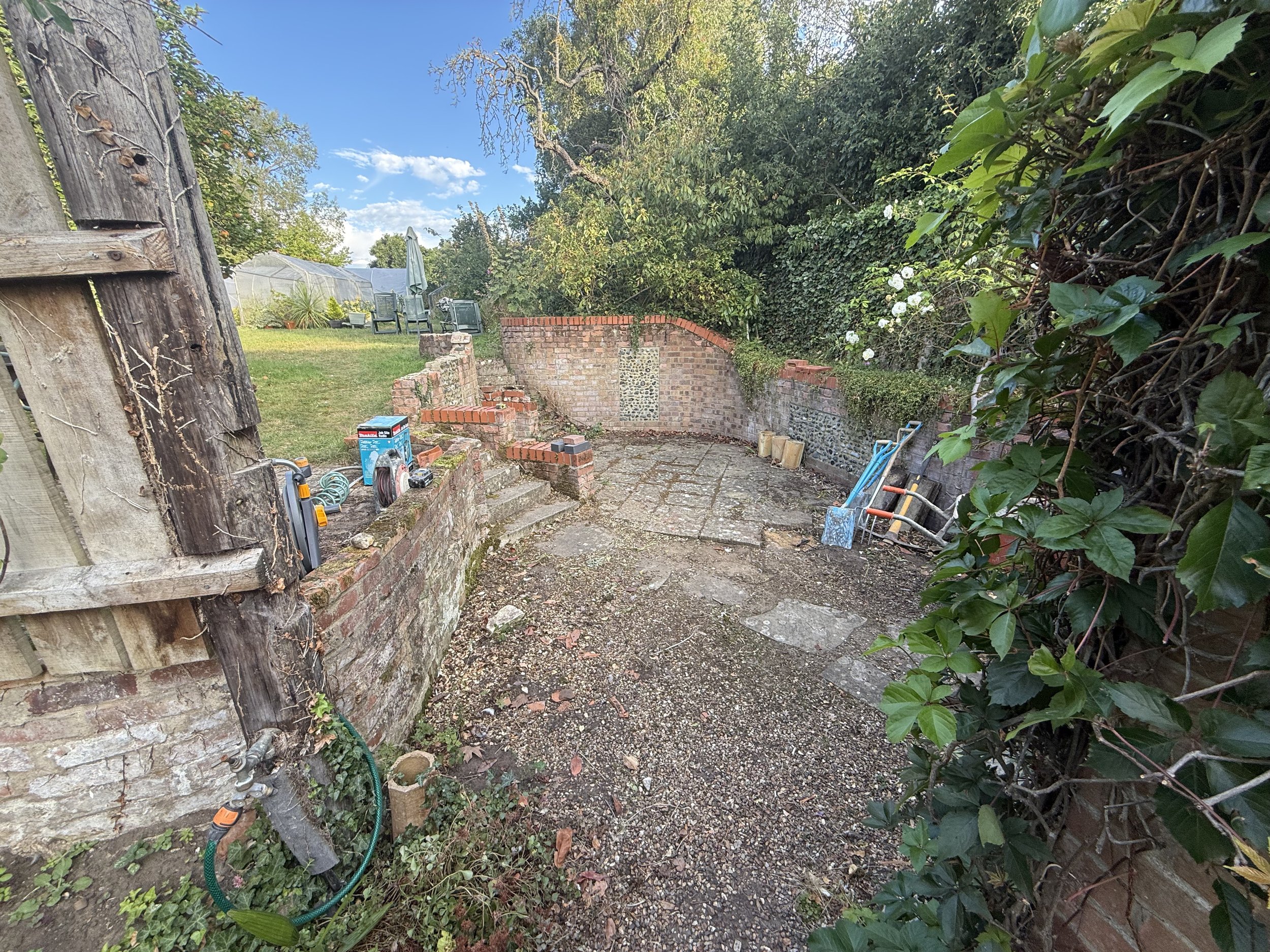 Backyard construction area with brick walls, gardening tools, and outdoor seating under a clear blue sky.