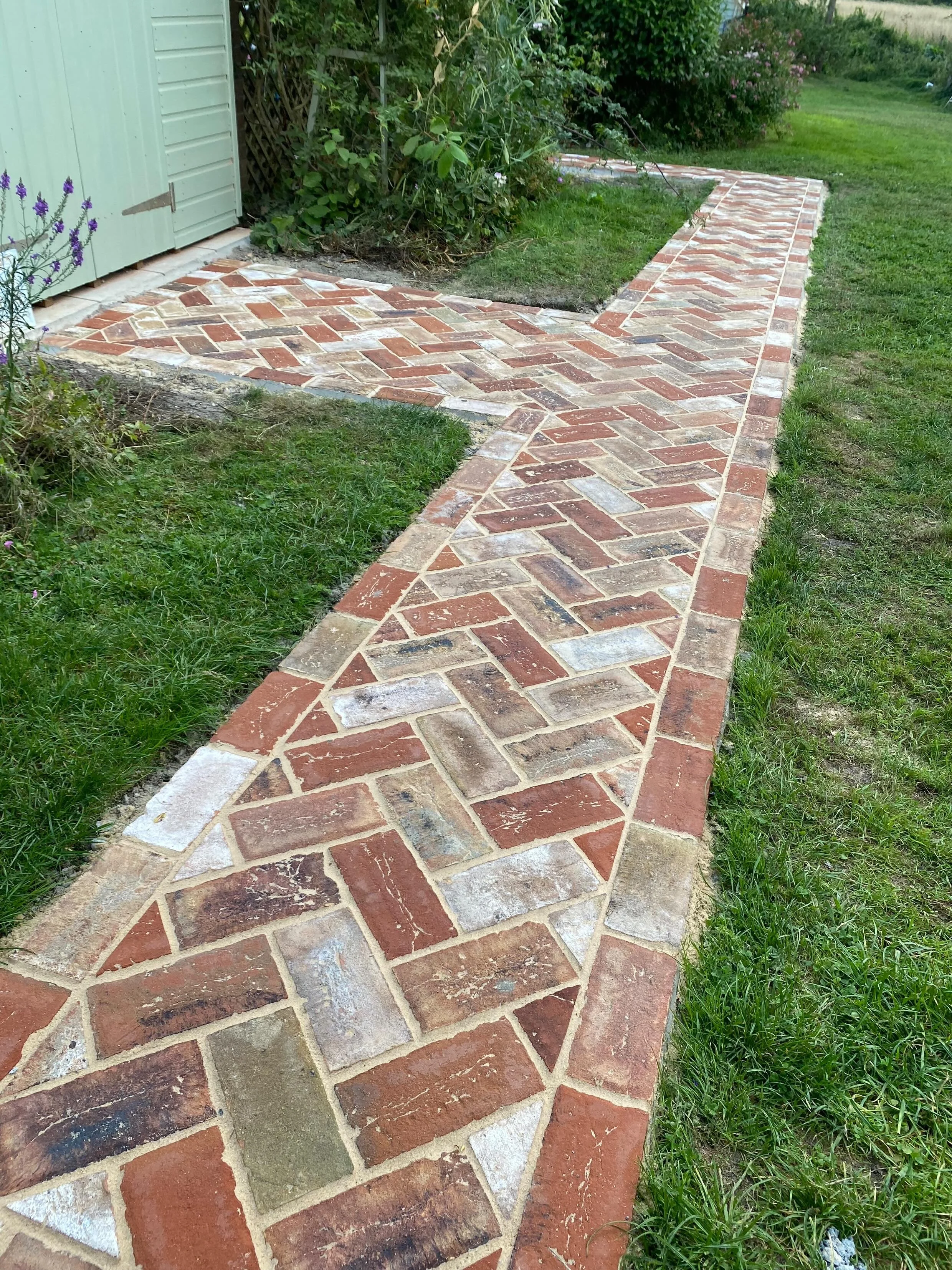 A brick pathway winding around a garden area with green grass and bushes, adjacent to a light blue building with closed shutters.