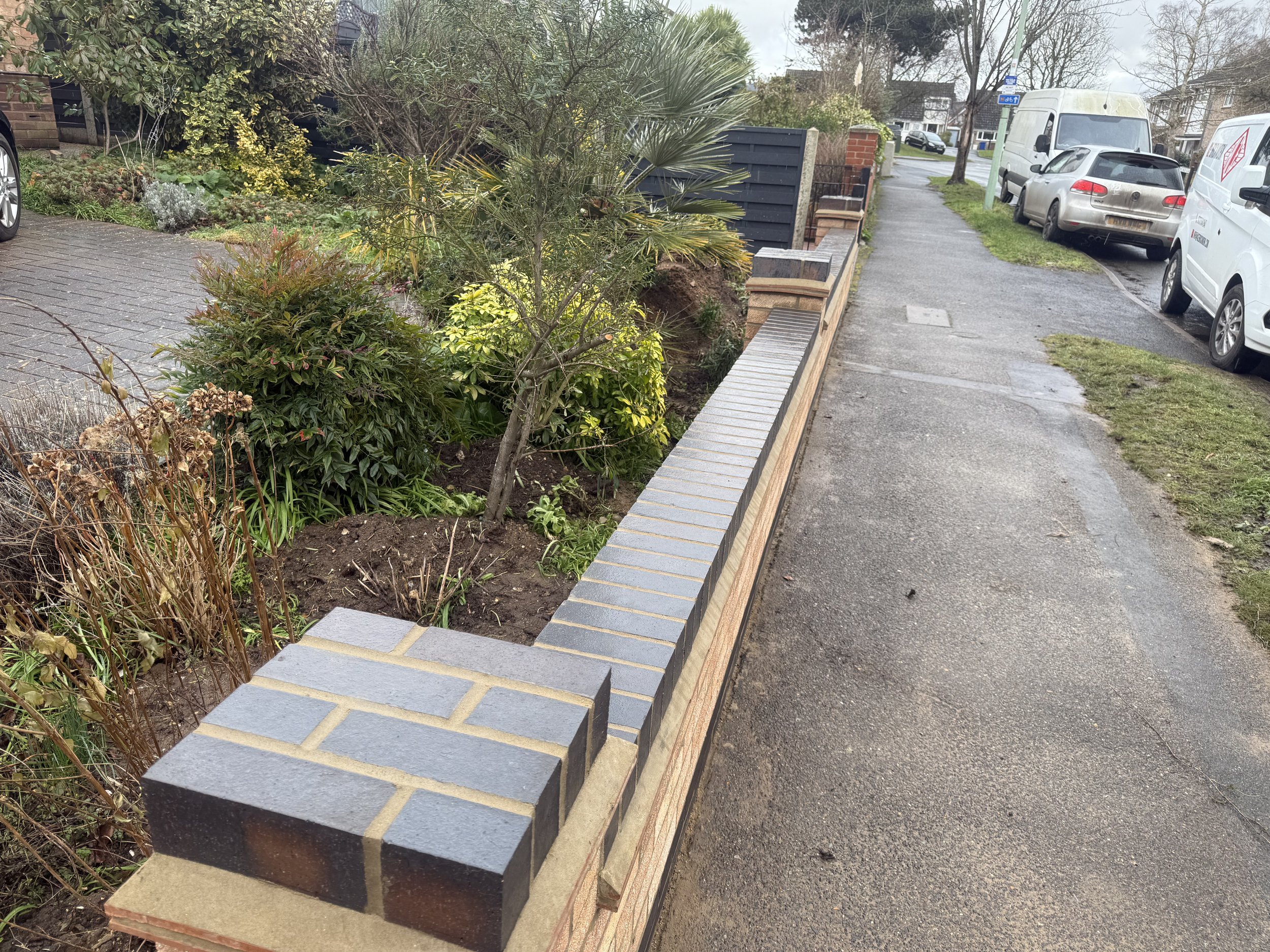 A sidewalk with a newly built brick wall separating it from a garden area with various plants, bushes, and small trees. Several parked cars and vans are visible along the street, with residential houses in the background.