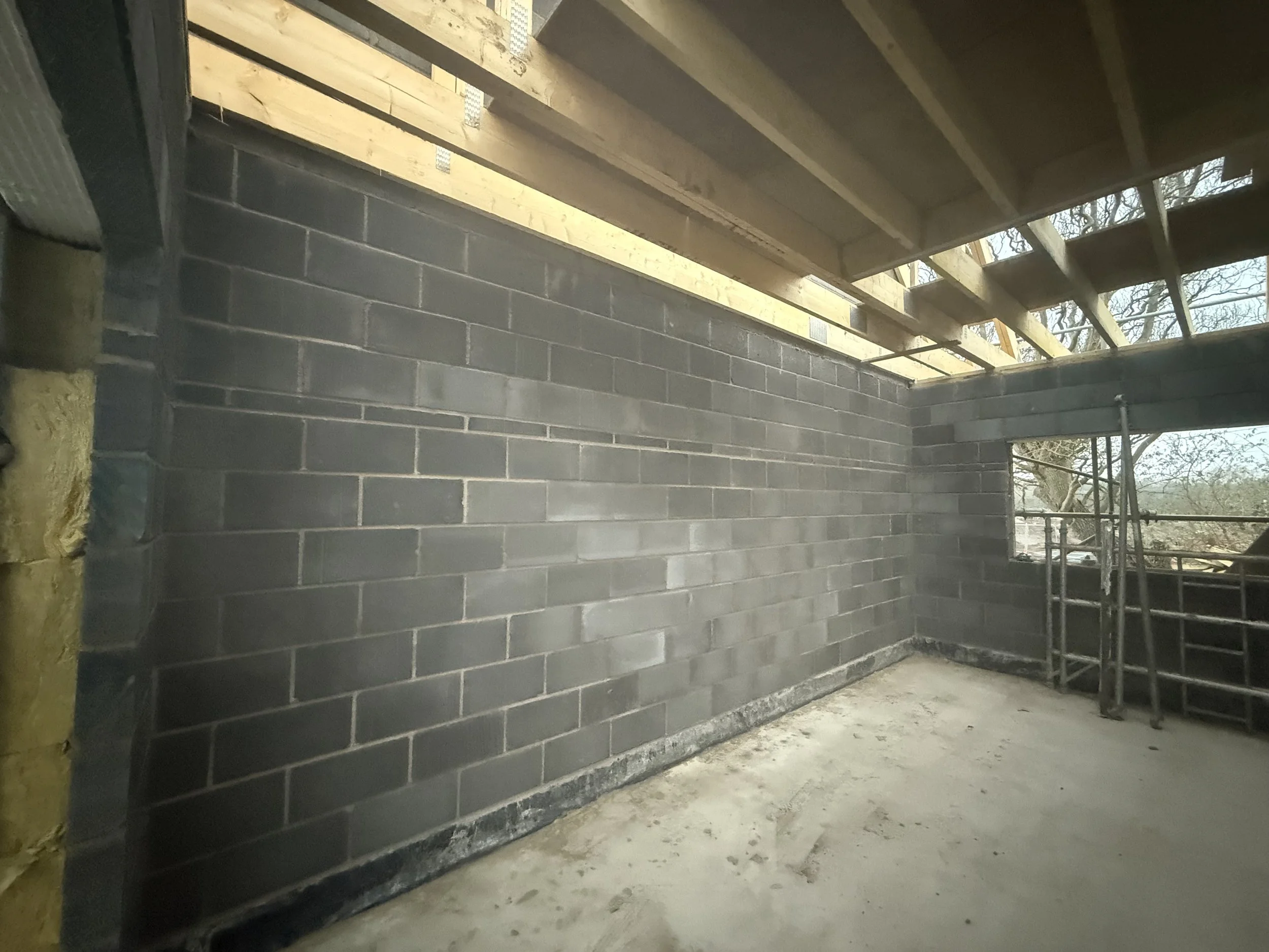 Interior view of a building under construction with a black brick wall, a window opening, and a wooden roof structure with scaffolding.