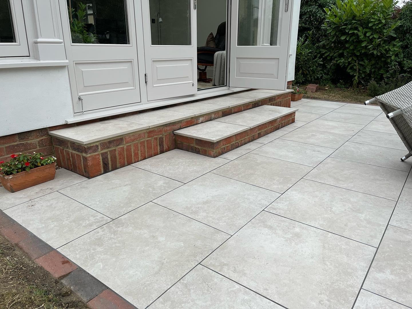 Residential patio with large beige tiles, brick steps leading to a sliding glass door, potted plants on the sides, and a wicker chair on the right.