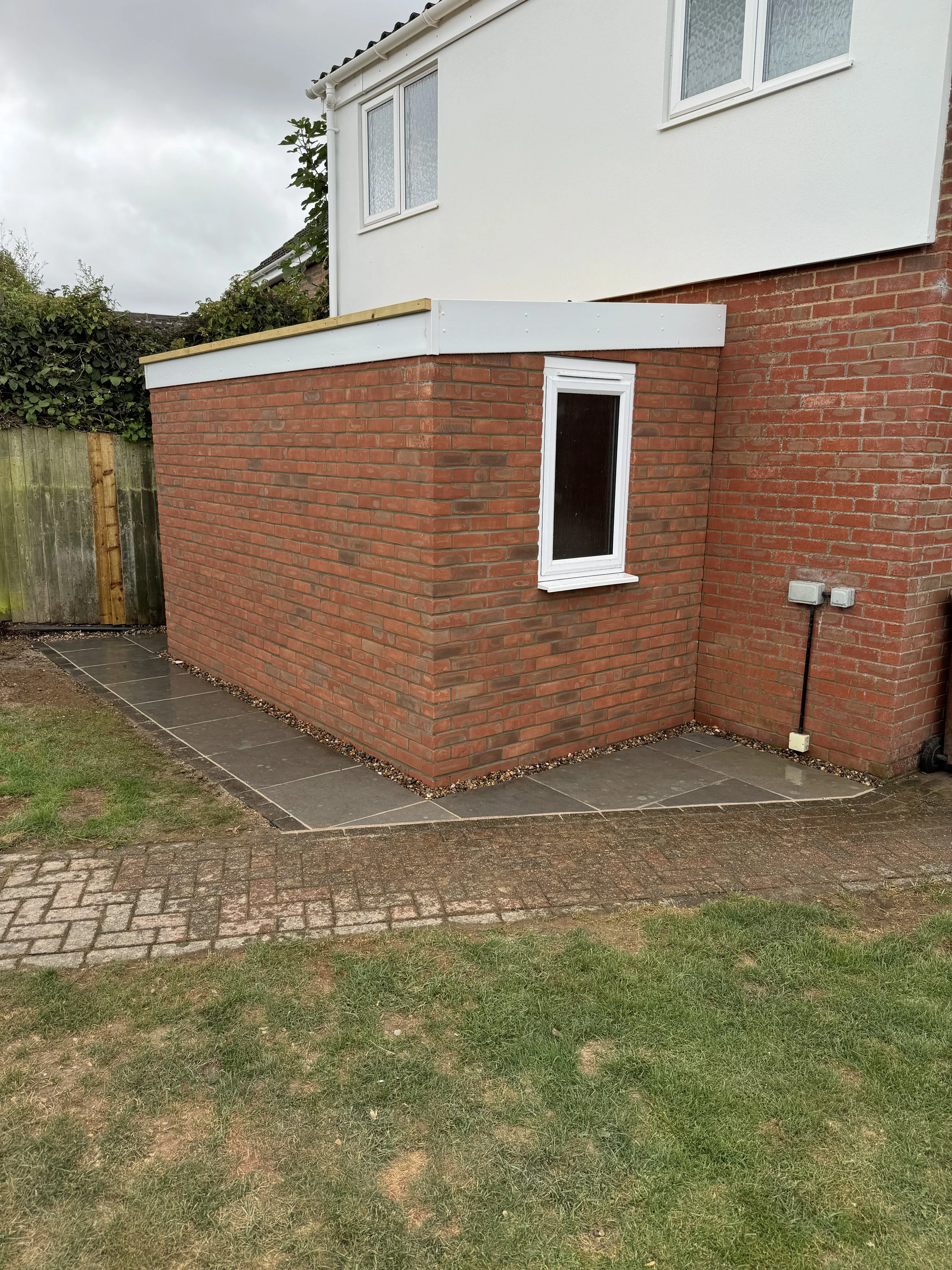 Back view of a brick extension on a house with a small window and new roofing, surrounded by a lawn, paving slabs, and a wooden fence.