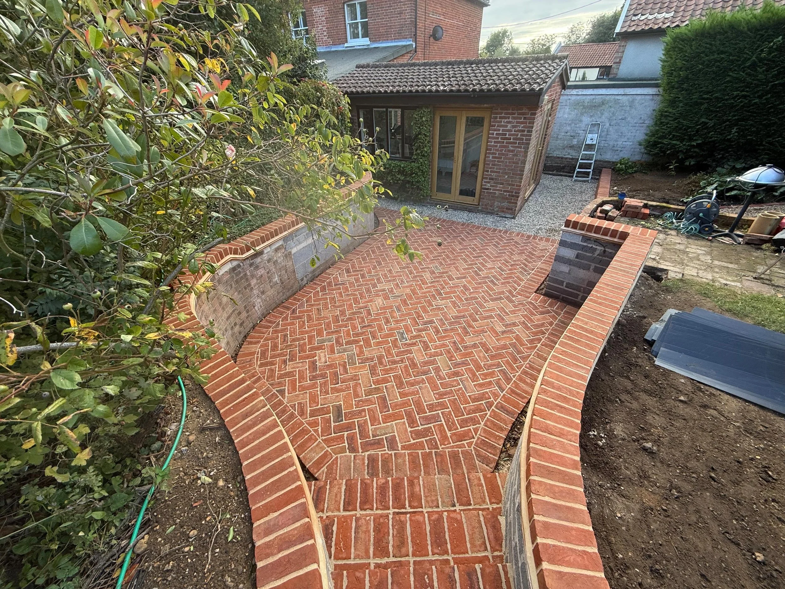 A newly constructed brick patio with a curved design, enclosed by brick walls, and a small brick shed in the background. The patio has a herringbone pattern, and there are construction tools and materials nearby.