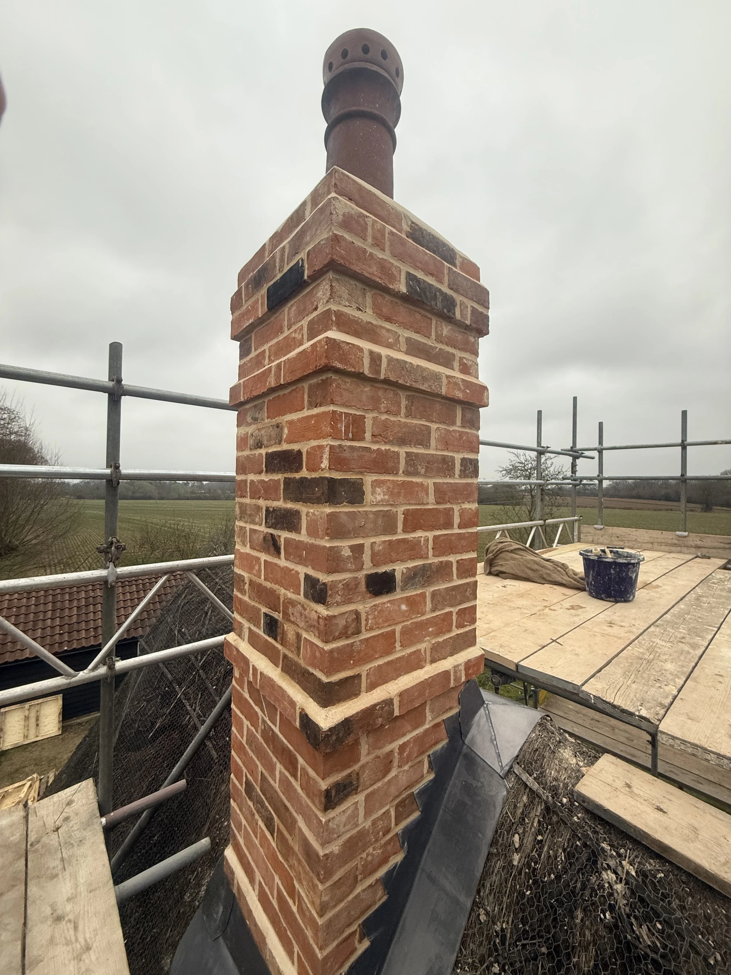A brick chimney with a metal cap on a roof under construction, with scaffolding and cloudy sky in the background.