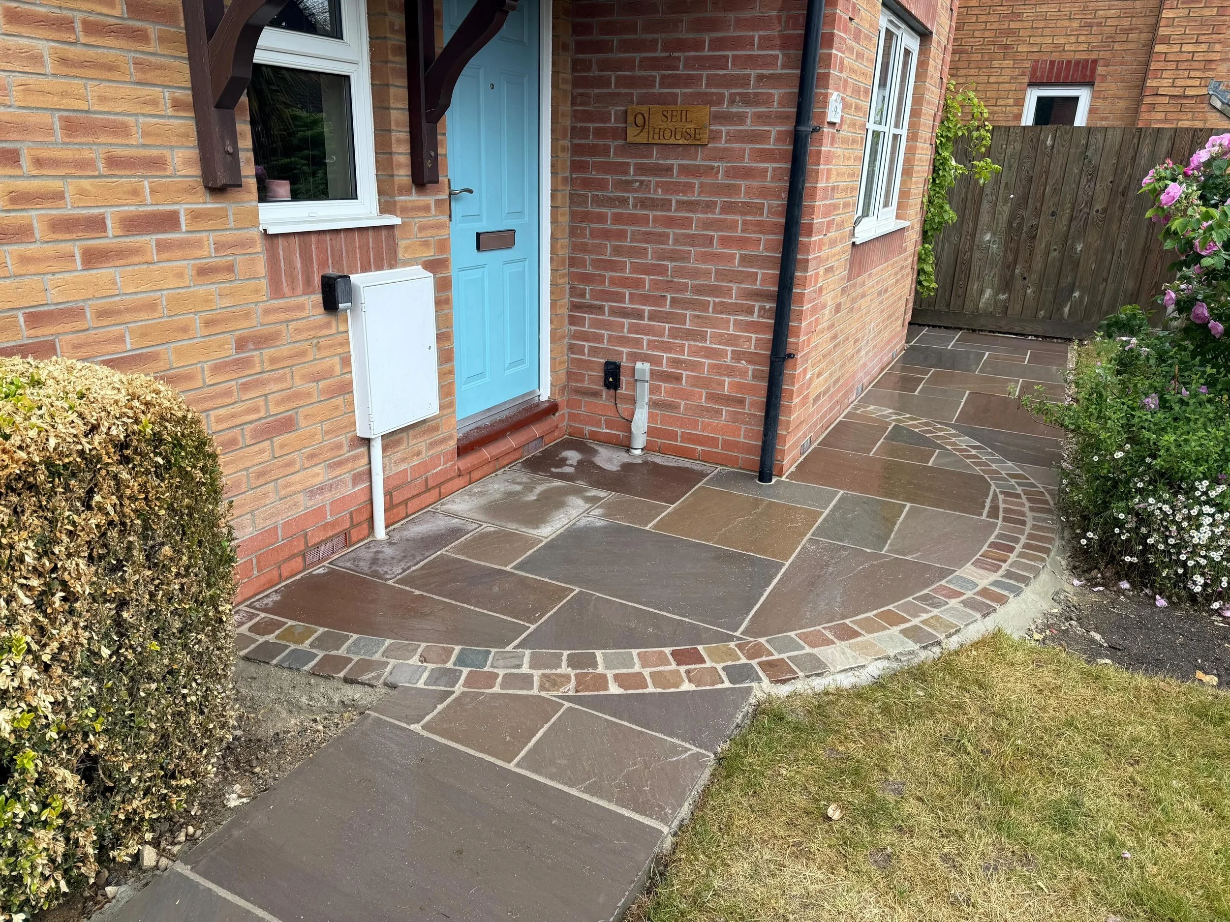 The front entrance of a brick house with a light blue door, a small window on each side, a wooden sign reading '9 SEIL HOUSE,' and a circular patio with multicolored stone paving and a small garden with flowers.