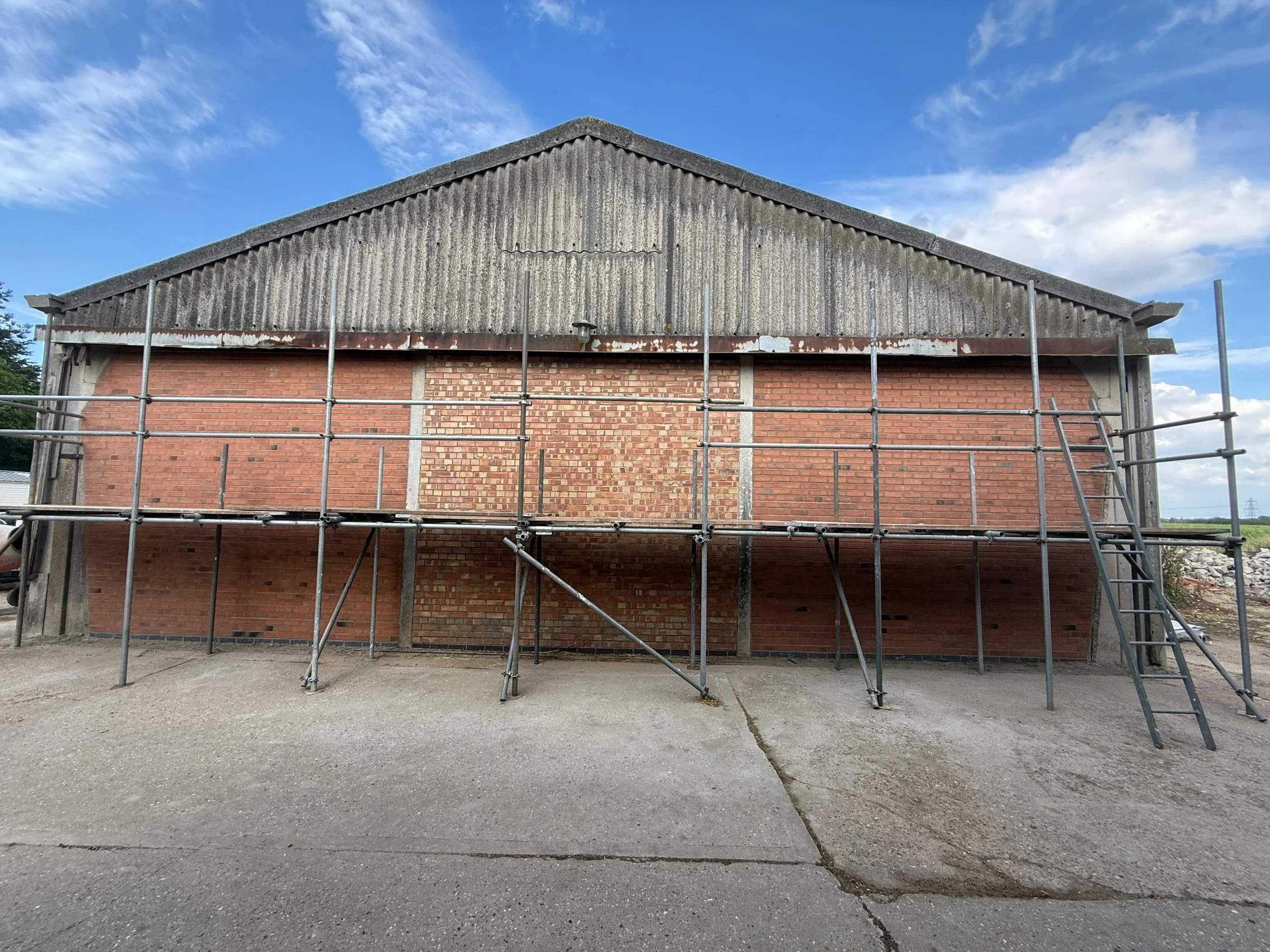 A building under construction with a brick facade, scaffolding around it, and a blue sky with clouds overhead.