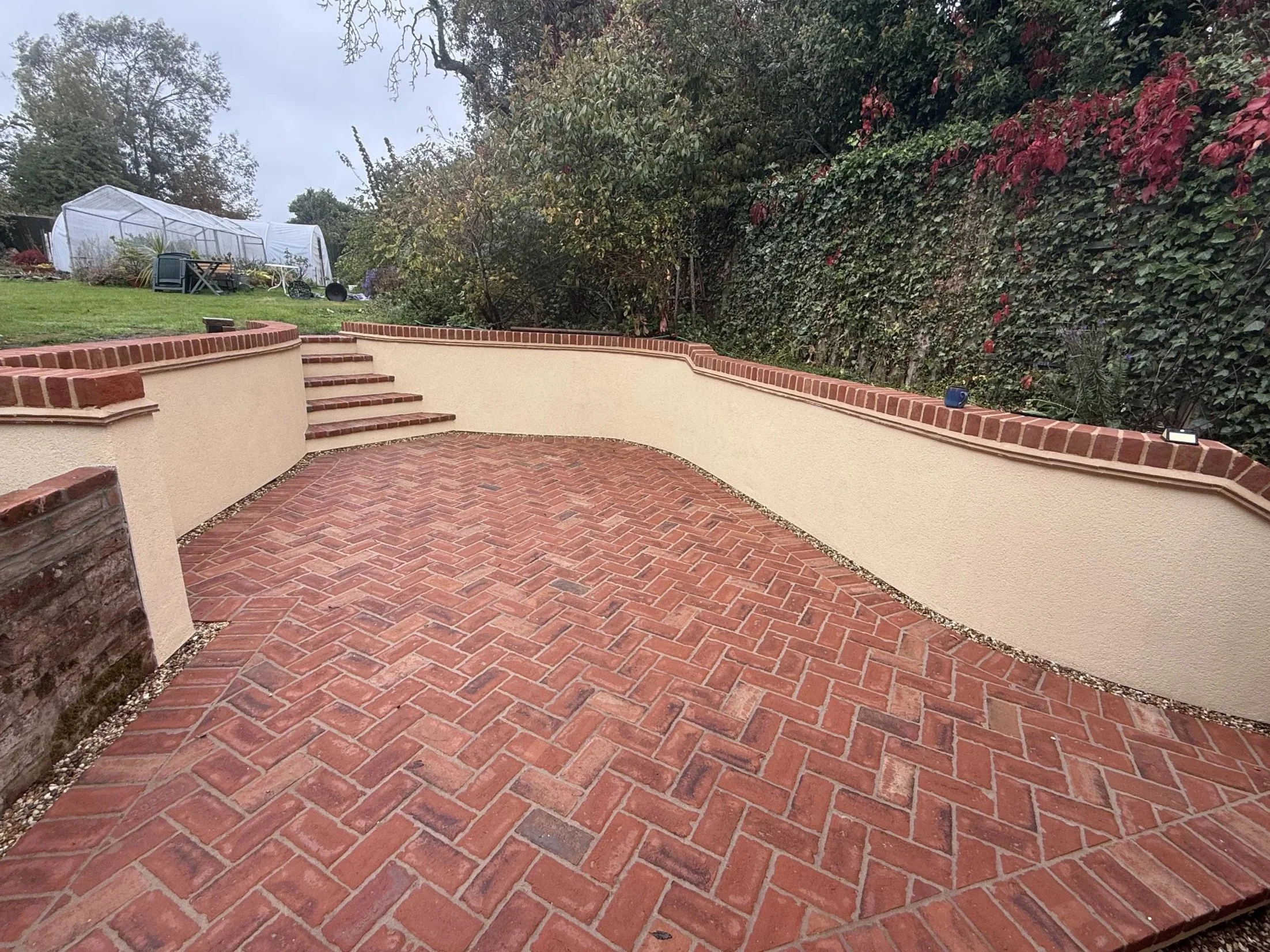 Freshly paved red brick patio with a brick and beige stucco retaining wall, outdoor garden area, and lush greenery in the background.