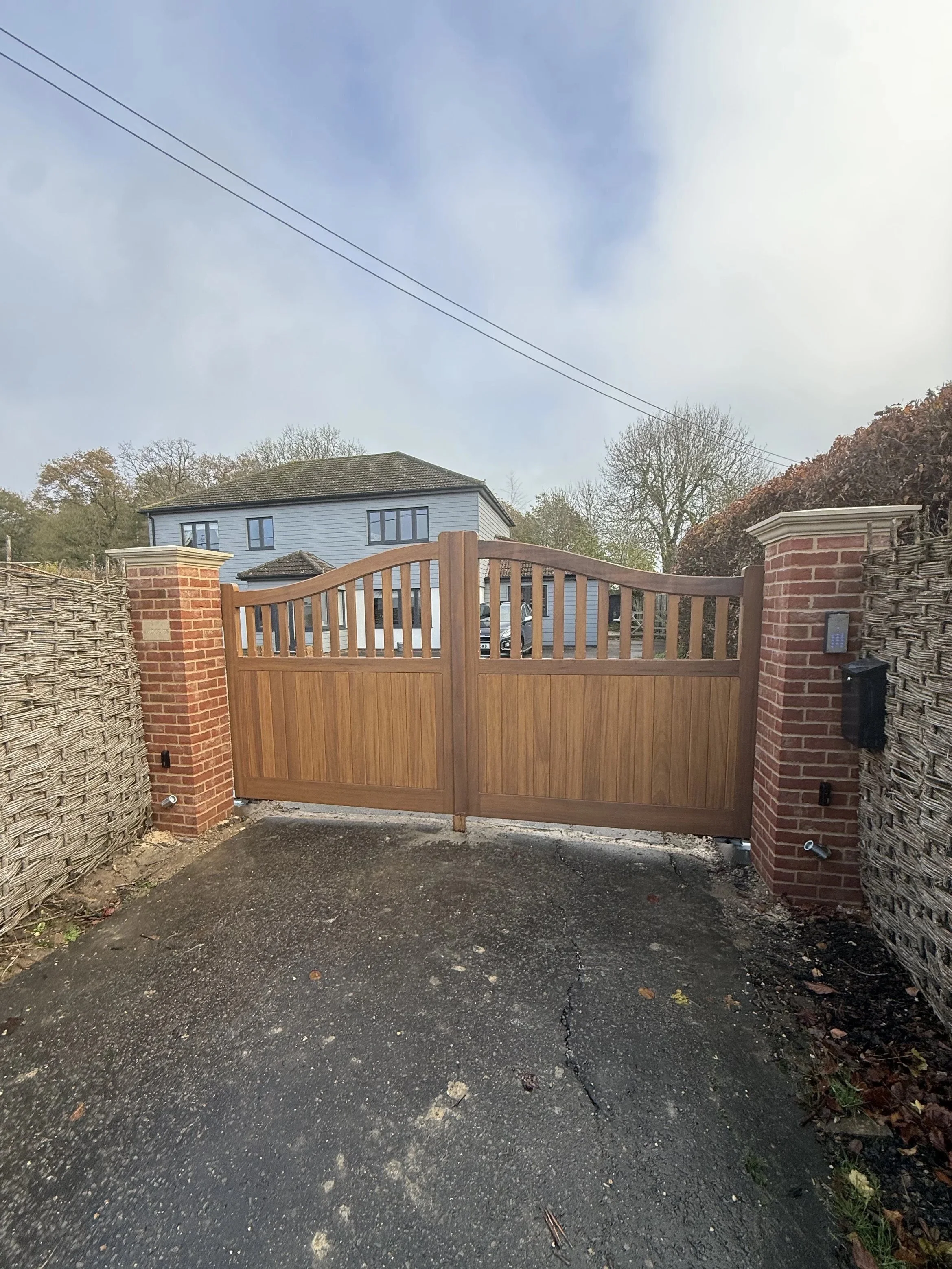 Wooden gate with brick pillars at entrance of a driveway, with a house and trees in the background.