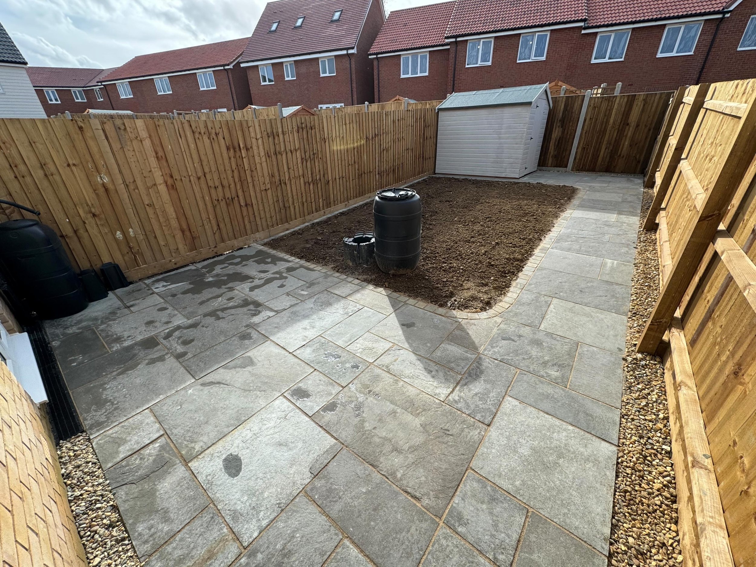 Partially paved backyard with stone tiles, a small area of bare soil, a wooden fence, garden shed, black water tank, and a small flower bed.