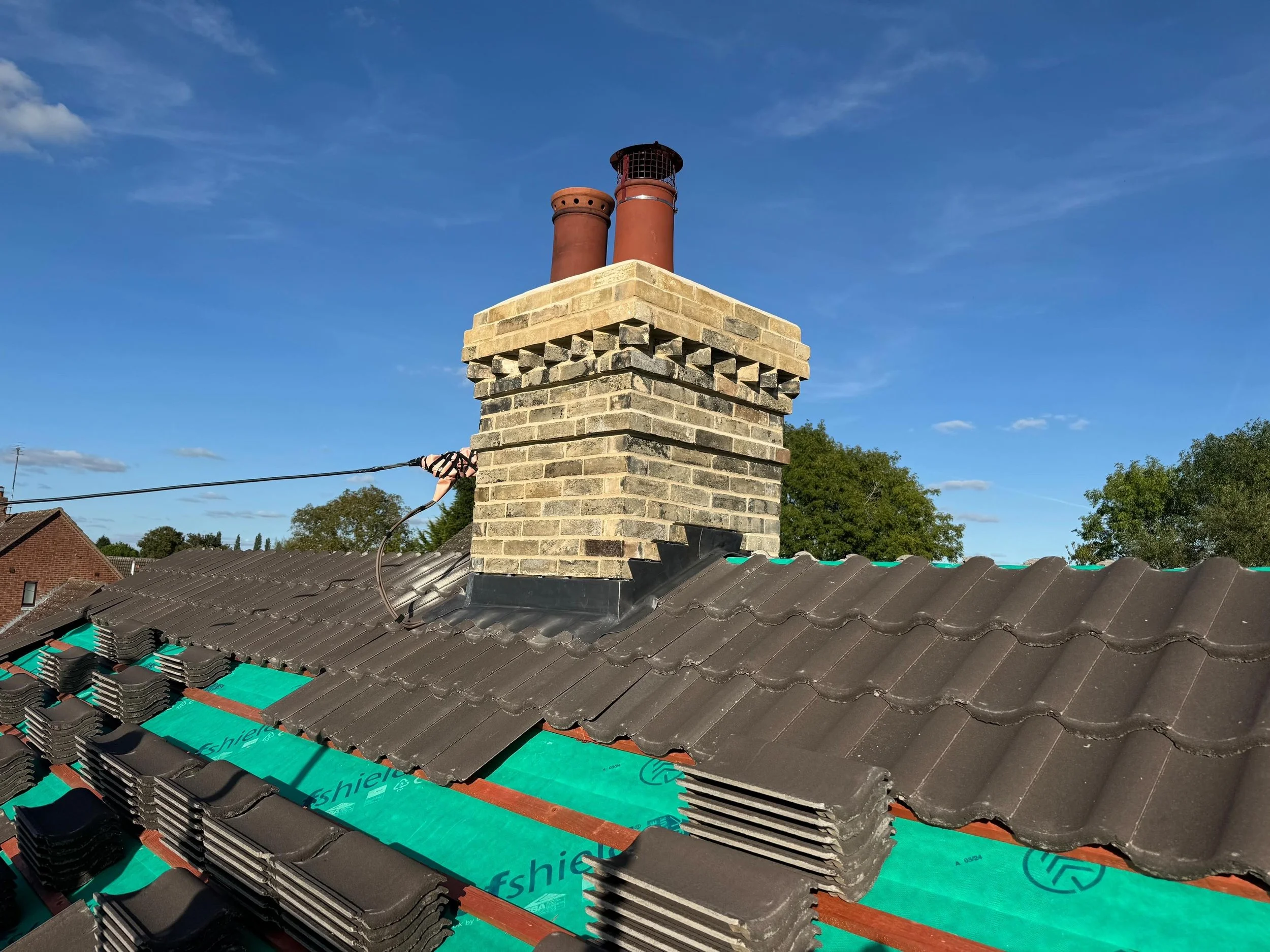Roof with new and old dark gray tiles, under construction, with a brick chimney and two red metal pipes against a clear blue sky and some trees in the background.