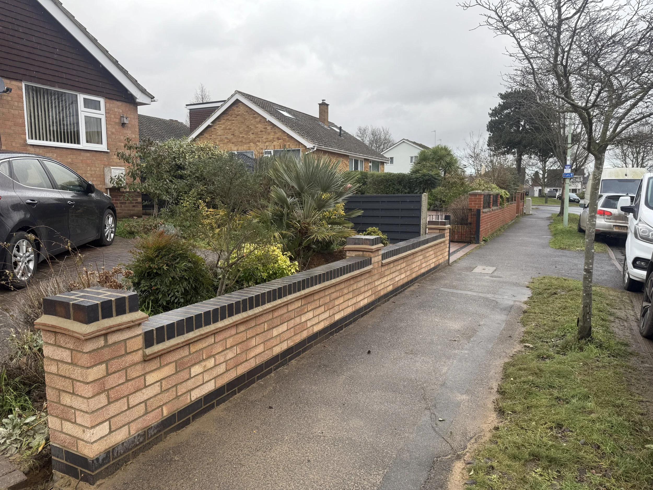 Residential sidewalk with a brick wall separating a garden and driveway, parked cars, and trees along the street under an overcast sky.