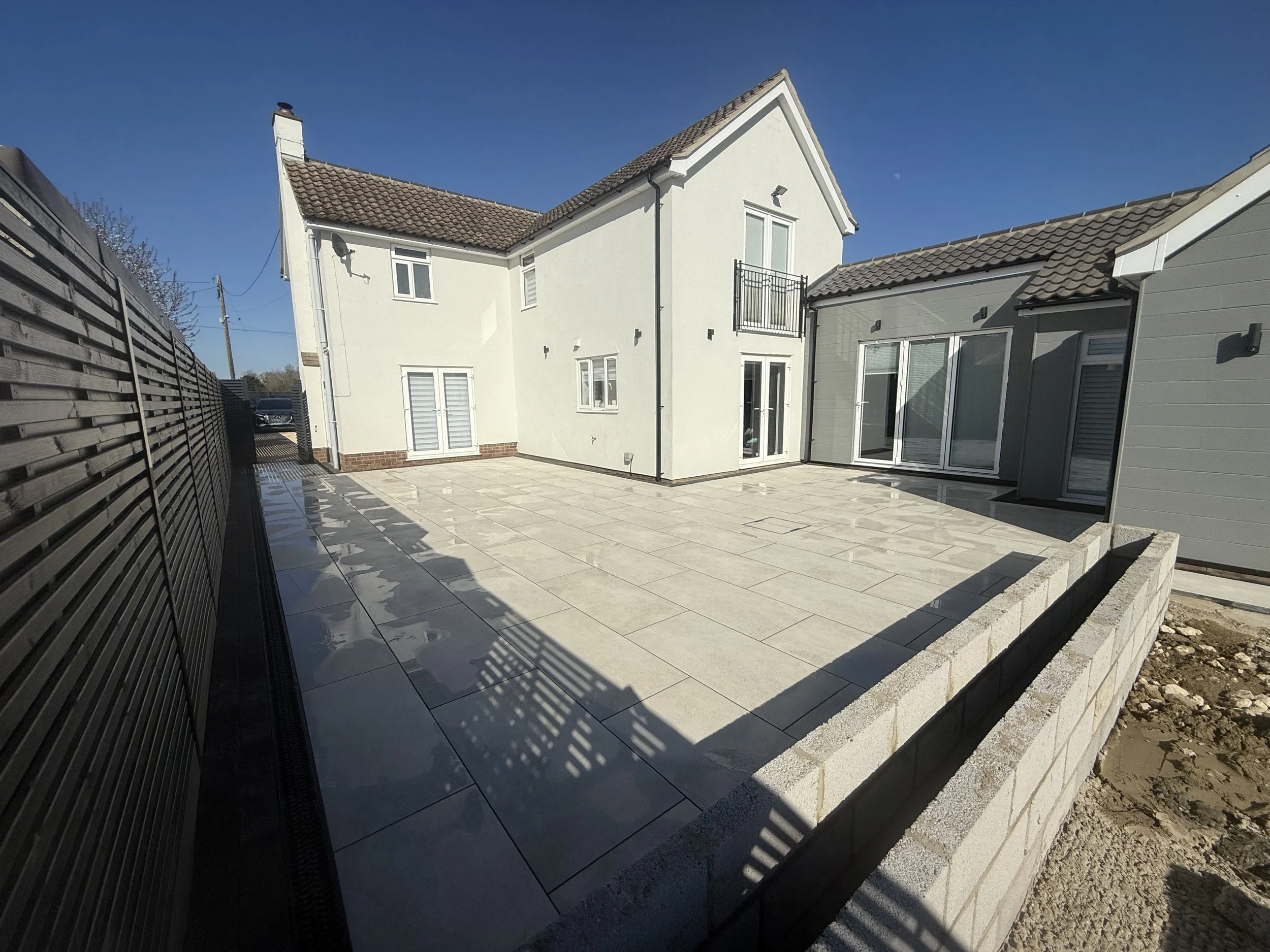 Newly paved outdoor patio with large concrete tiles, enclosed by a low brick wall, adjacent to a modern white house with large windows and a small balcony, under a clear blue sky.