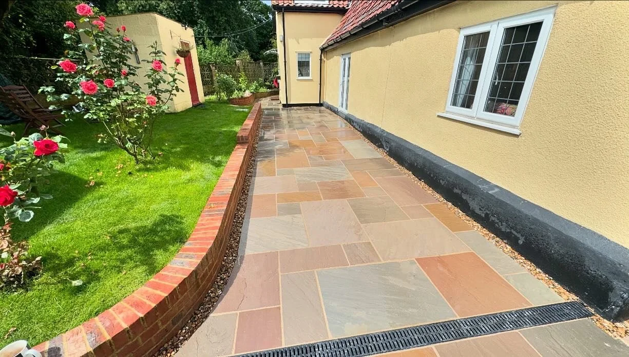 A paved patio area alongside a yellow house with white-framed windows, next to a garden with green grass, a rose bush with pink flowers, and a small yellow shed.