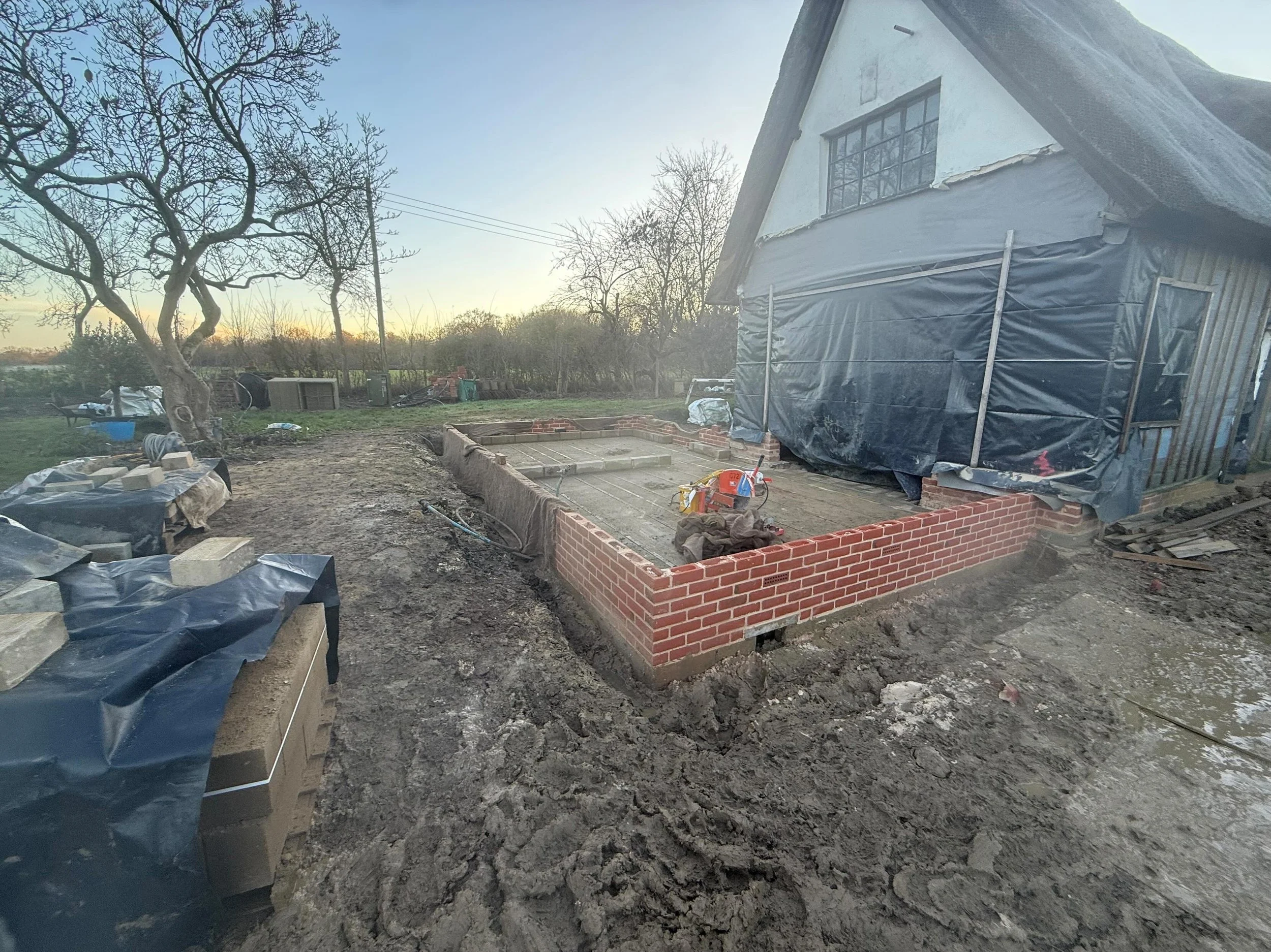 Construction site around a house with partially built brick foundation, construction materials, and tools, during sunset.