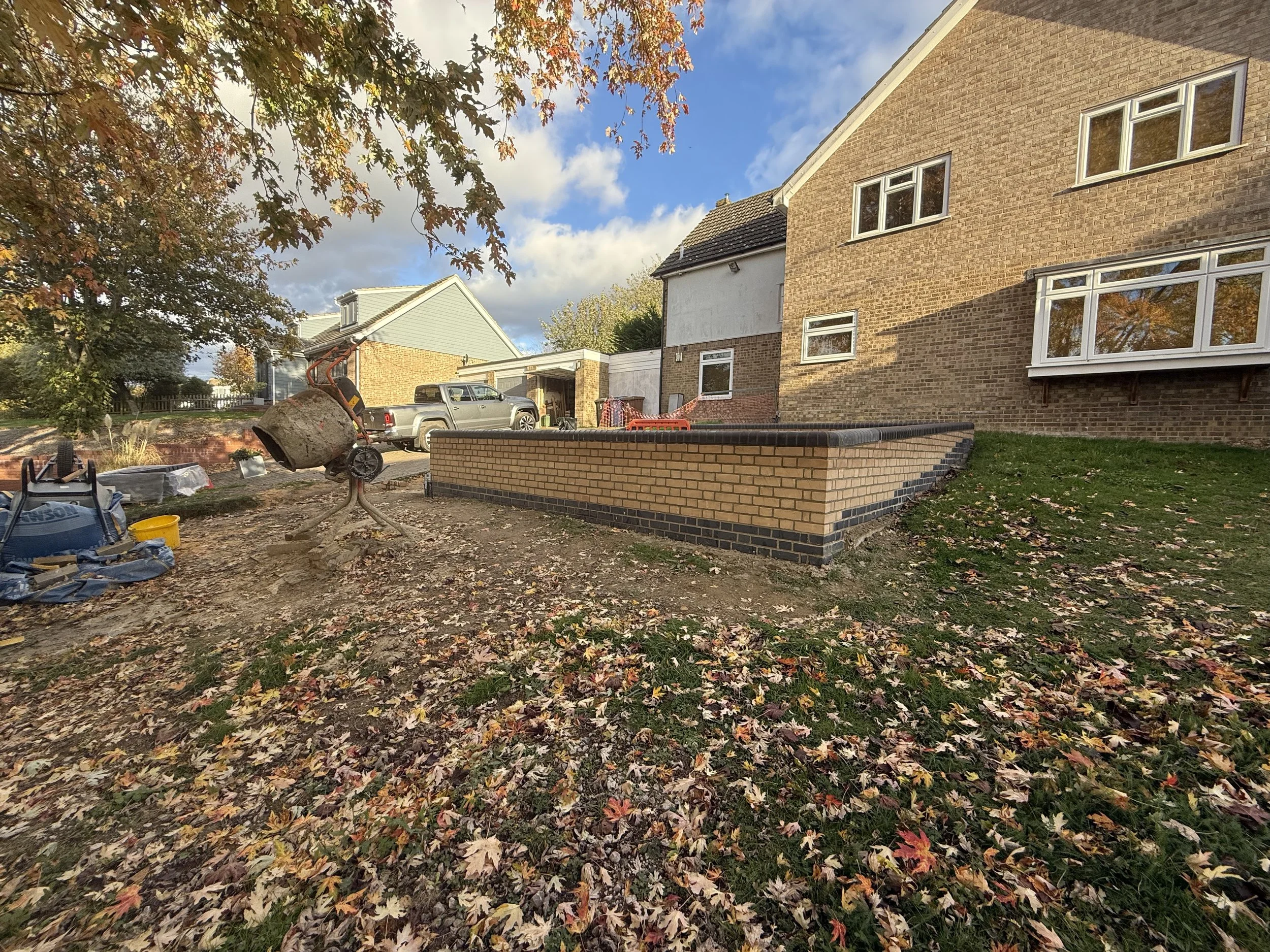A residential backyard with a partially finished brick retaining wall, construction tools, a cement mixer, and fallen autumn leaves on the ground under a partly cloudy sky.