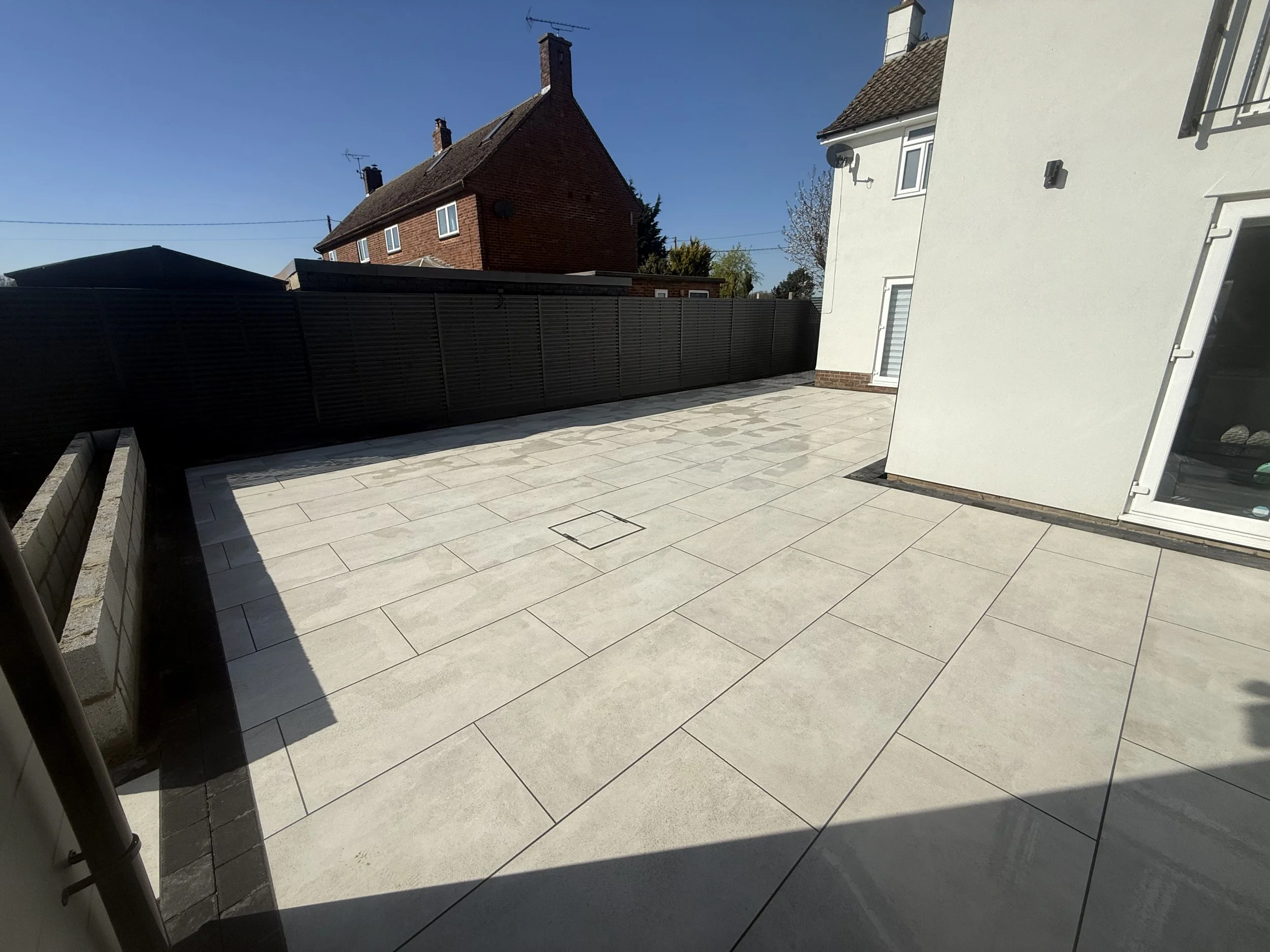 Empty outdoor patio with large rectangular light-colored tiles, a black fence, a white house wall, and neighboring brick houses in the background on a sunny day.